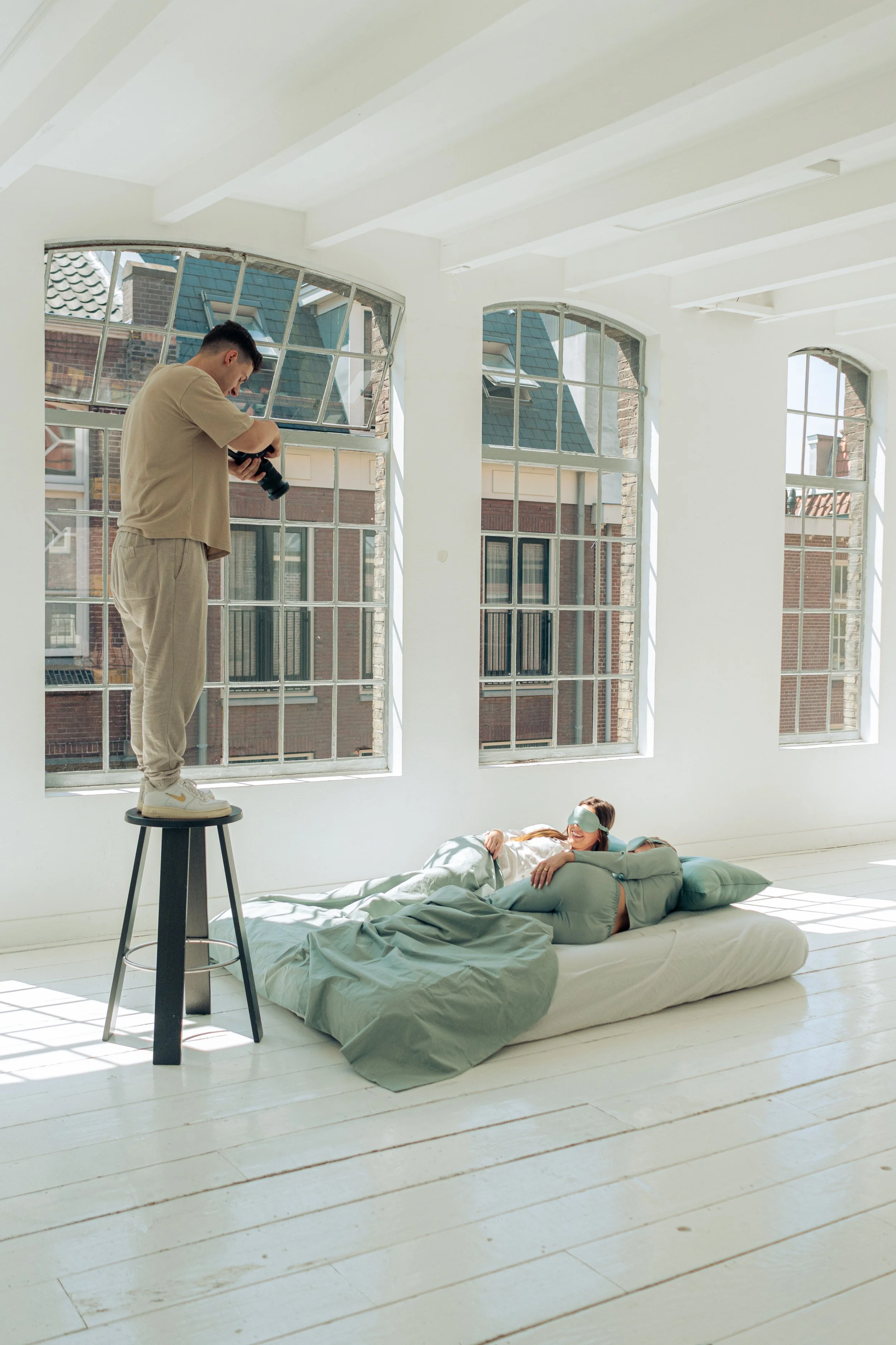 Een man staat op een krukje en neemt een foto van een vrouw die op een matras ligt, terwijl ze een slaapmasker draagt in een lichte kamer met grote ramen.