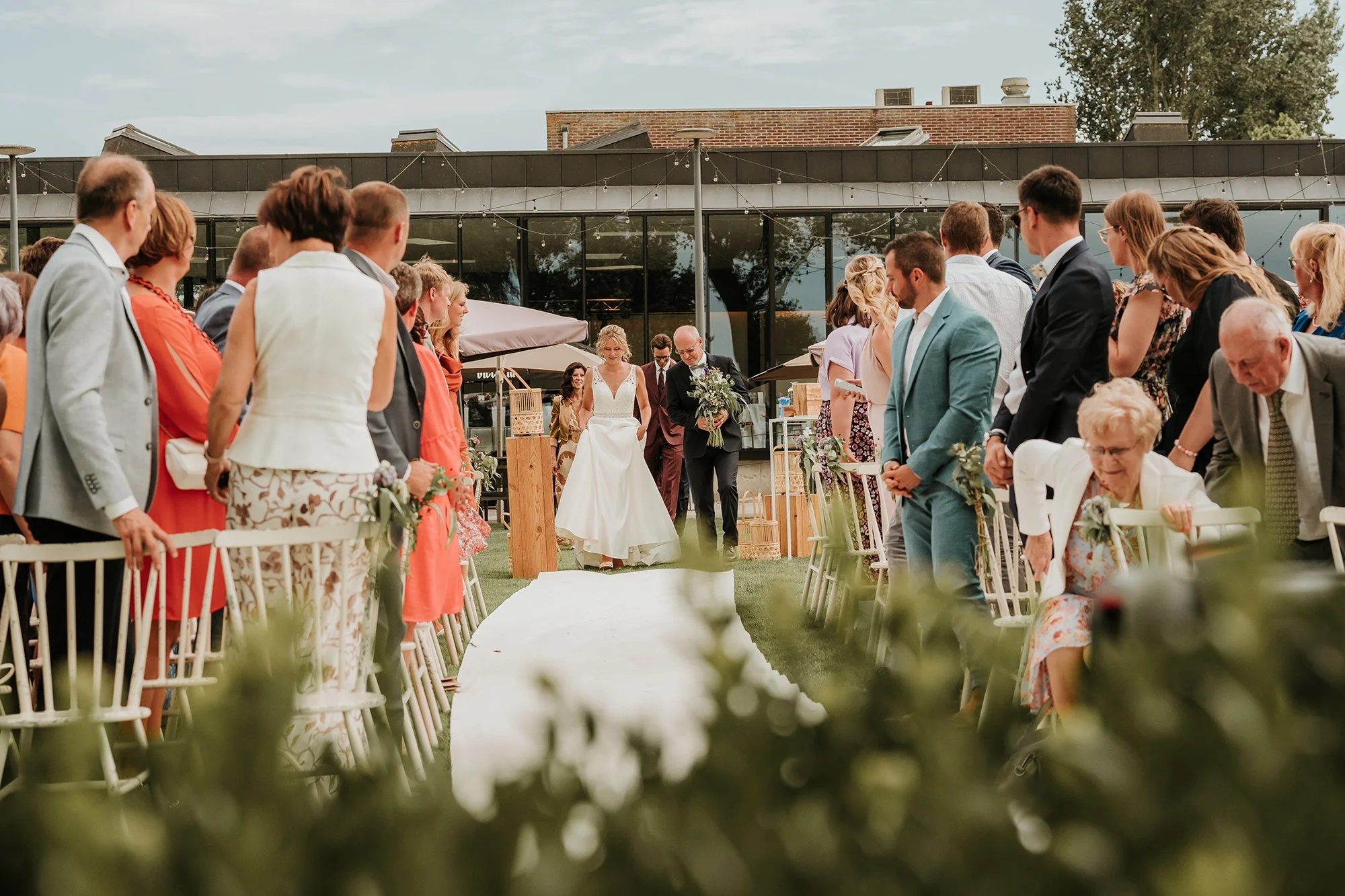 Gelnokt huwelijk op een buitenterras met gasten die staan en zitten, kerkelijke plechtigheid met bruid en bruidegom in het midden, onder een glazen overkapping, met een moderne gebouw op de achtergrond.