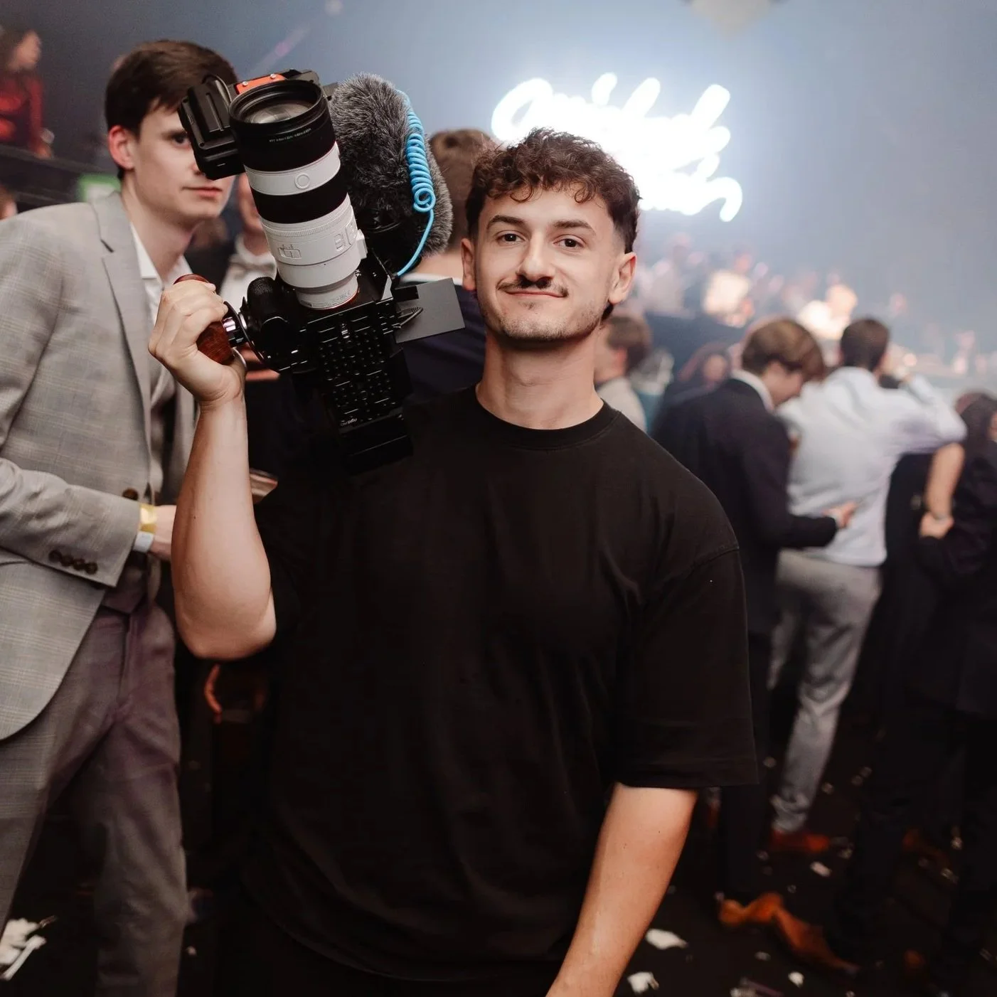 Man met korte, curly haar en een gestructureerde snor poseert met een professionele camera op een schouder tijdens een evenement, met een menigte mensen op de achtergrond.
