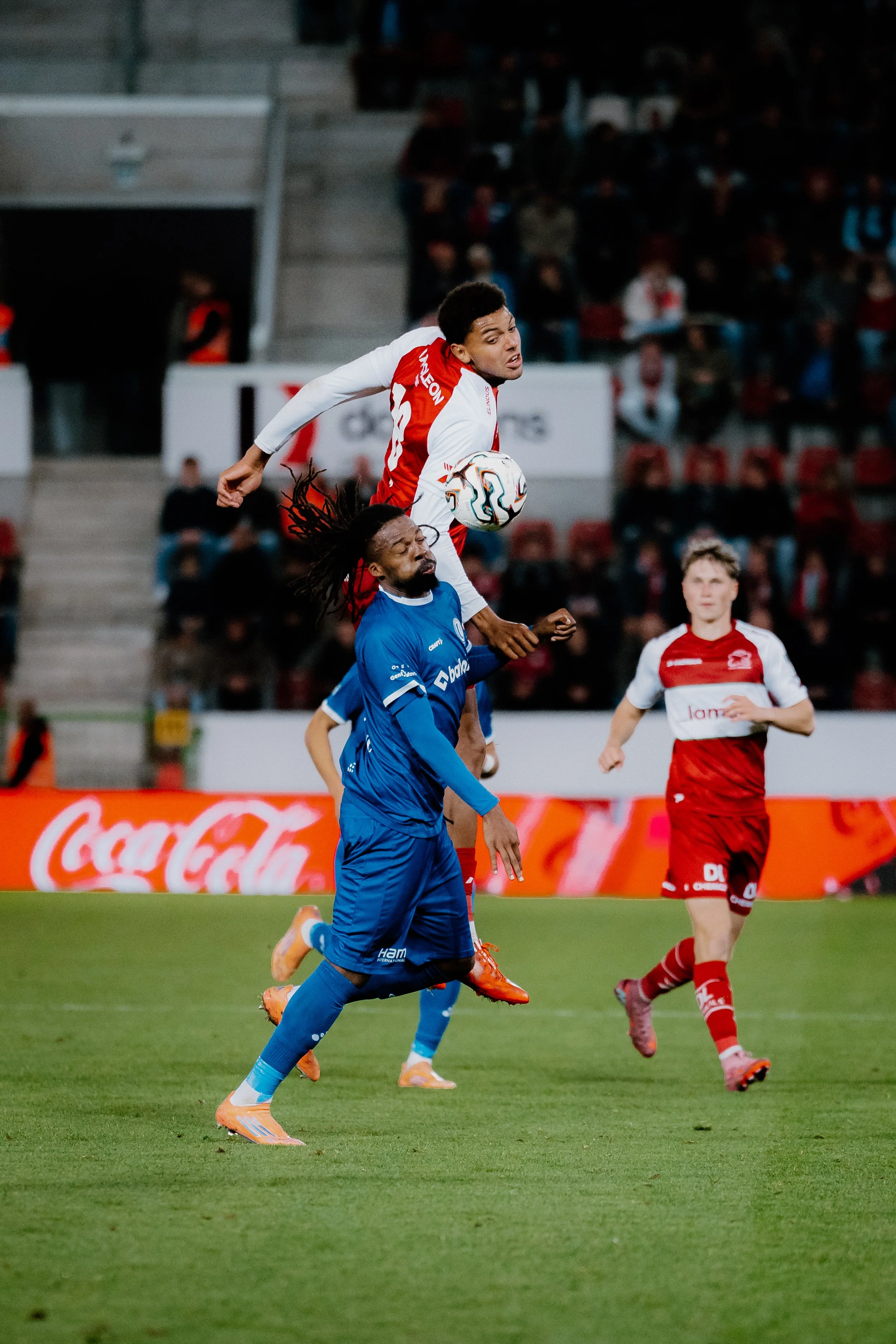 Een voetbalwedstrijd met een speler in een rood-wit tenue die springt en de bal in de lucht probeert te spelen, terwijl een tegenstander in een blauw tenue onder hem staat. Er zijn andere spelers en een publiek zichtbaar op de achtergrond.