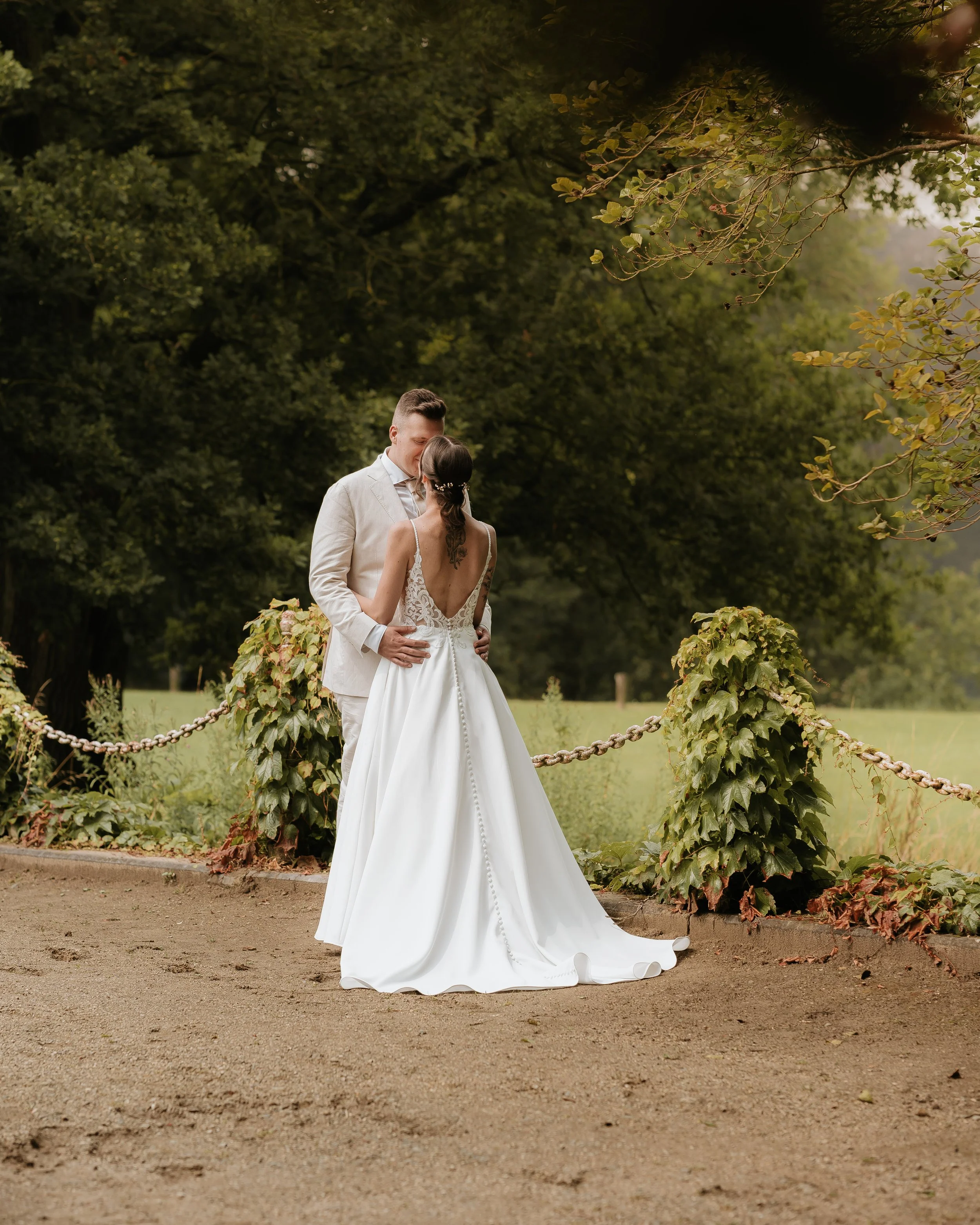 Een huwelijksfoto van een paar dat elkaar in een parkje kust, met groene bomen en een stenen pad.