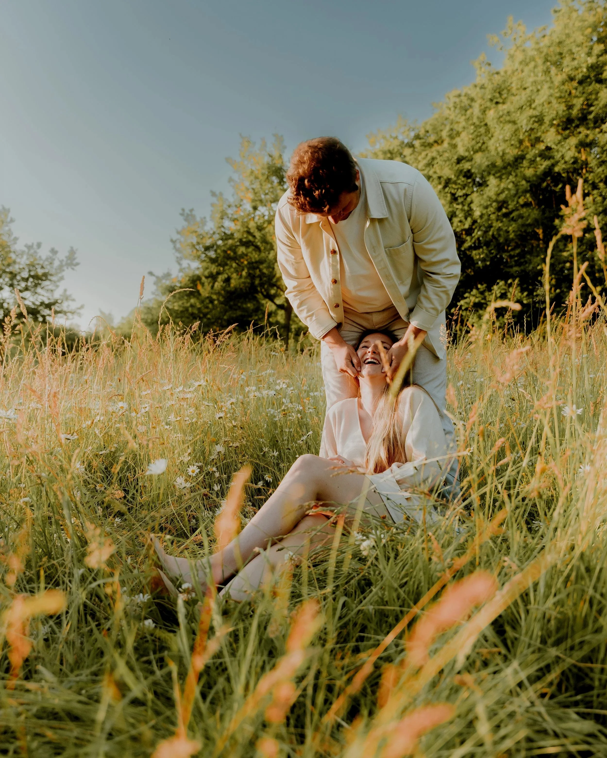 Twee mensen, een man en een vrouw, lachen samen in een veld met gras en bloemen, met bomen op de achtergrond en een blauwe hemel boven hen.