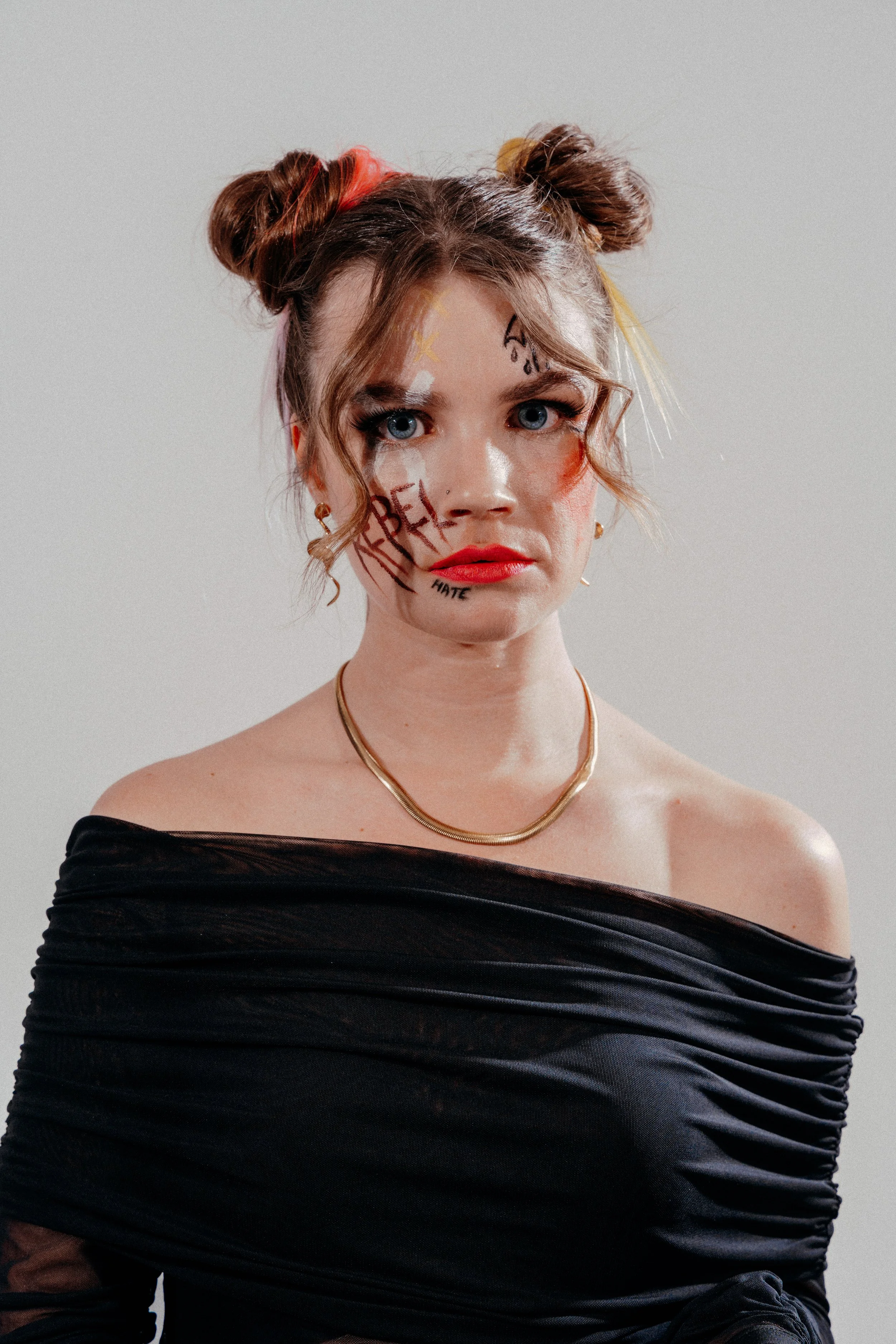 Portret van een jonge vrouw met twee kleurrijke haarstijlen, opvallende make-up, woorden en woorden op haar gezicht, en een zwarte off-shoulder top, tegen een neutrale achtergrond.