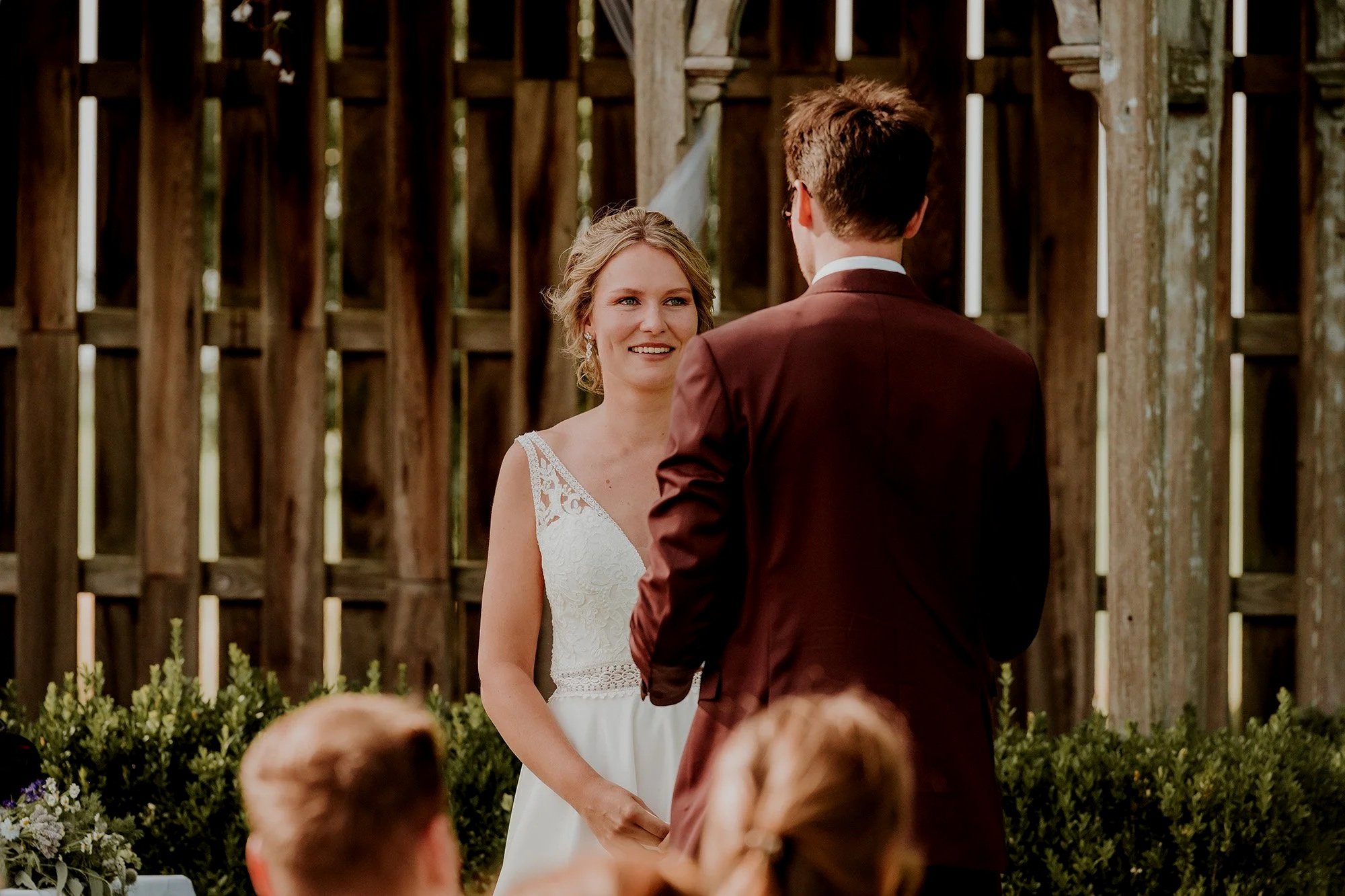 Bruidsseremonie met een bruid en een bruidegom die elkaar aankijken, buiten, met houten achtergrond en gasten.