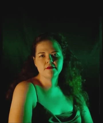 A woman with long, curly hair and red lipstick, illuminated with green lighting, looking at the camera against a dark background.