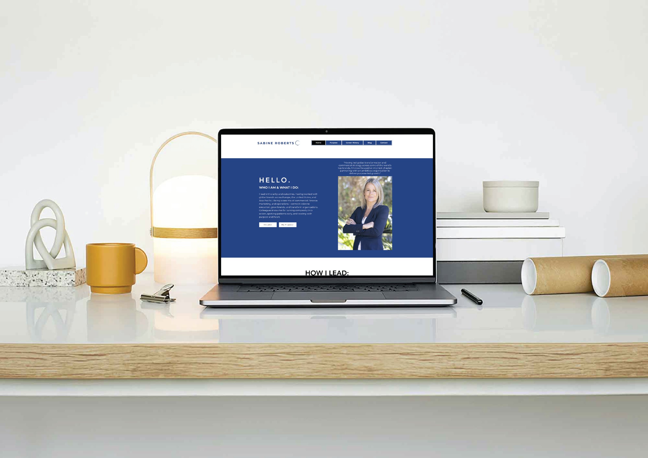 Open laptop on a white desk displaying a personal website with a blue background, a photo of a woman, and text including 'HELLO' and 'WHO I AM & WHAT I DO'. Surrounding the laptop are a yellow mug, a clip, a small white lamp, a stack of books with a white container on top, and cardboard tubes.