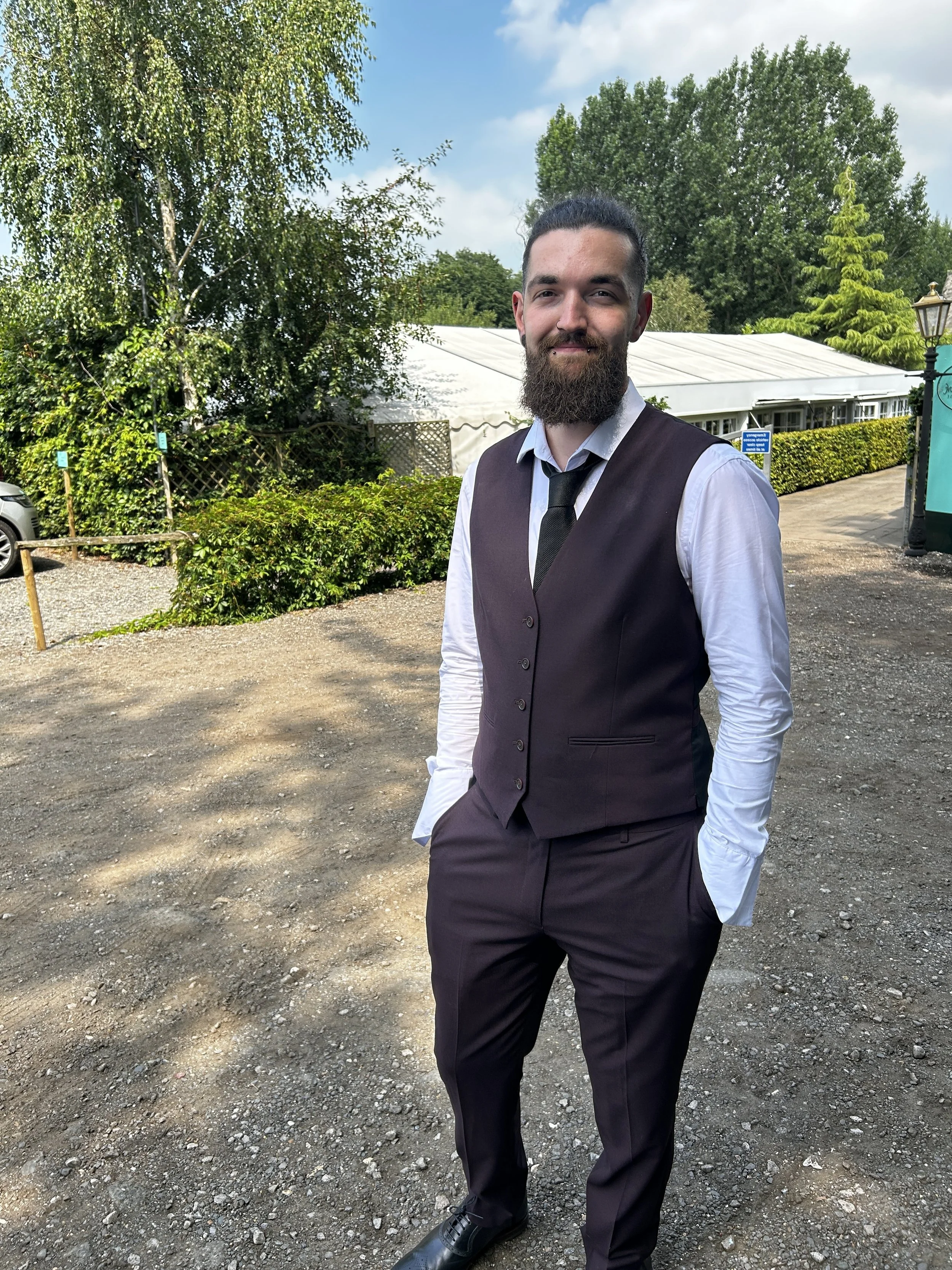 A man with a beard and mustache wearing a white dress shirt, dark vest, and tie, standing outdoors with trees and a white tent in the background.