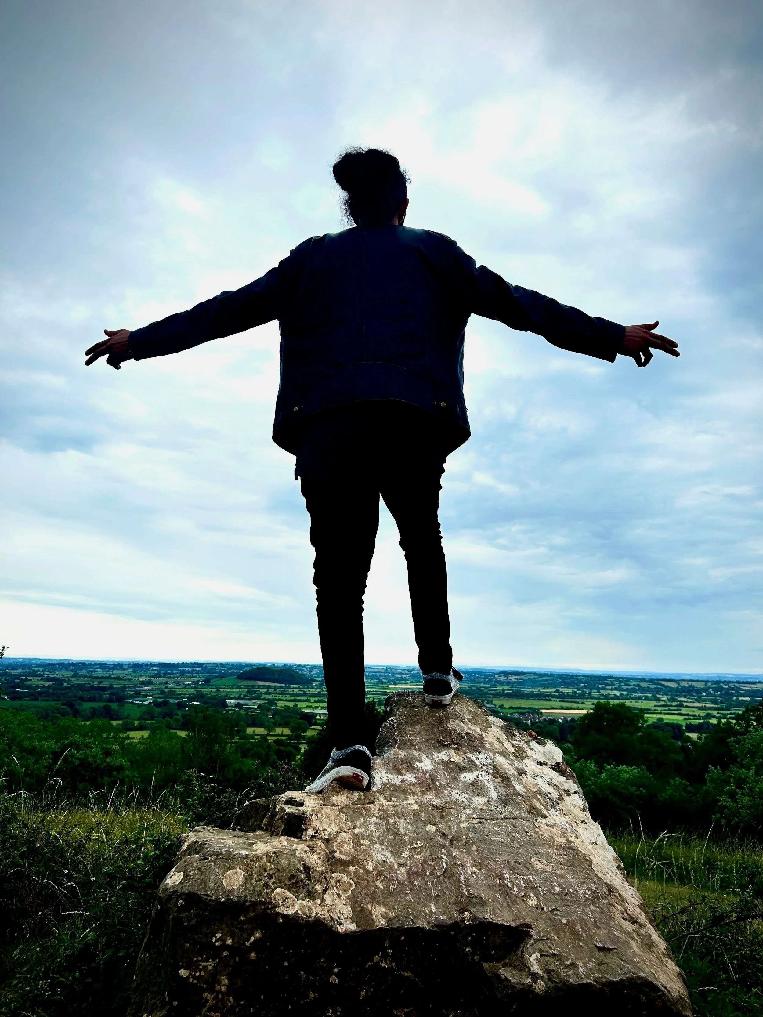Person standing on a large rock with arms stretched out, overlooking a vast green landscape with a cloudy sky.
