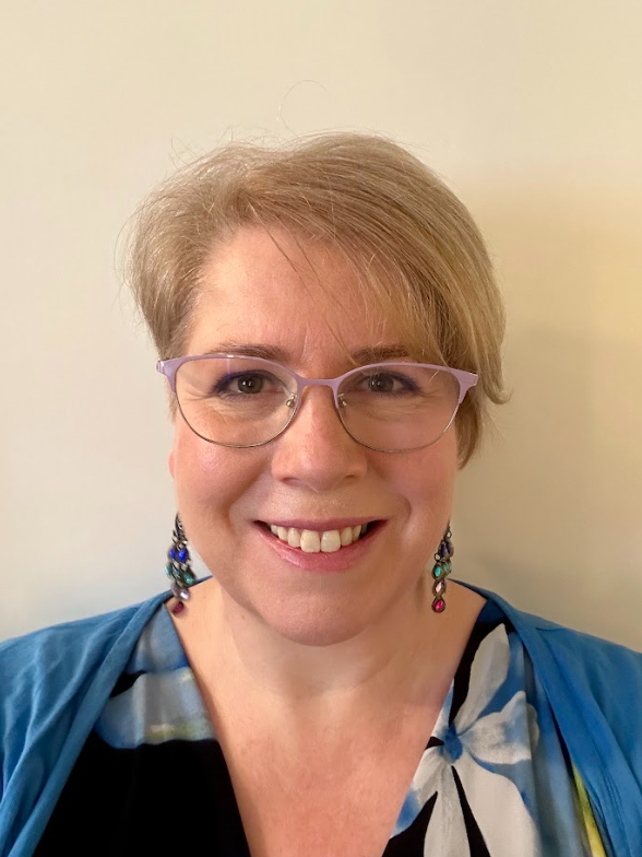 A woman with short blonde hair, purple glasses, and colorful earrings, smiling at the camera against a plain light-colored background.
