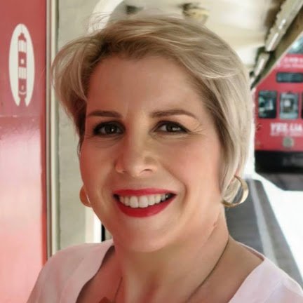Smiling woman with short blonde hair and red lipstick, standing outdoors near a bus and a red emergency telephone box.