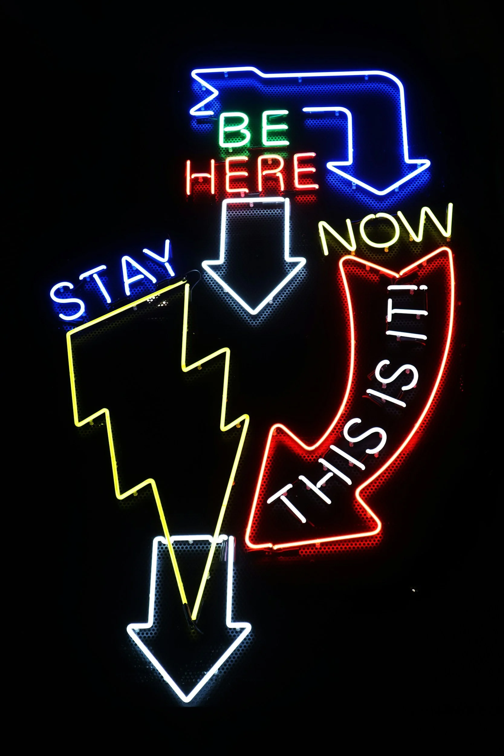 Neon sign with arrows and text reading 'Be Here Now', 'Stay', 'This Is It', with arrows pointing down and to the right. The sign is colorful with blue, green, red, yellow, and white neon lights.