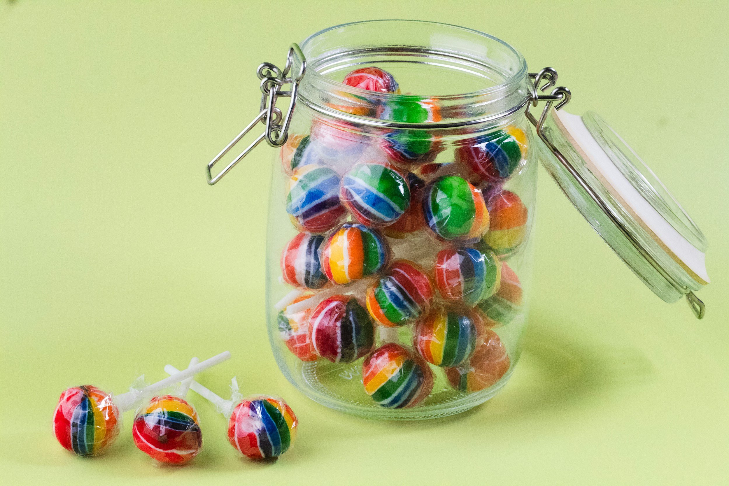 A glass jar filled with rainbow-striped lollipops, some outside the jar on a light green surface.
