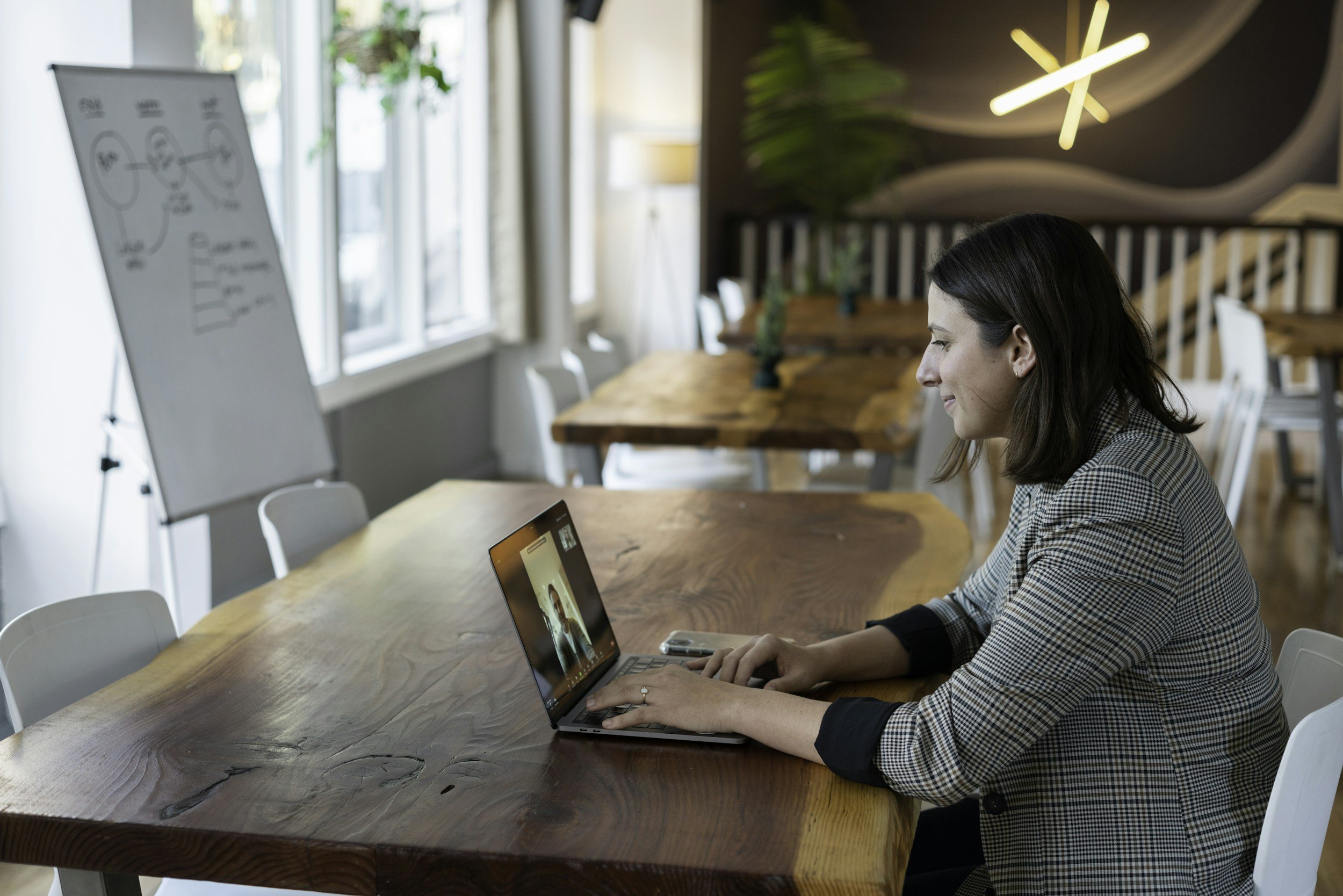 Woman sitting at a wooden table, engaging in a video call on her laptop, in a bright, modern conference room with large windows and plants.