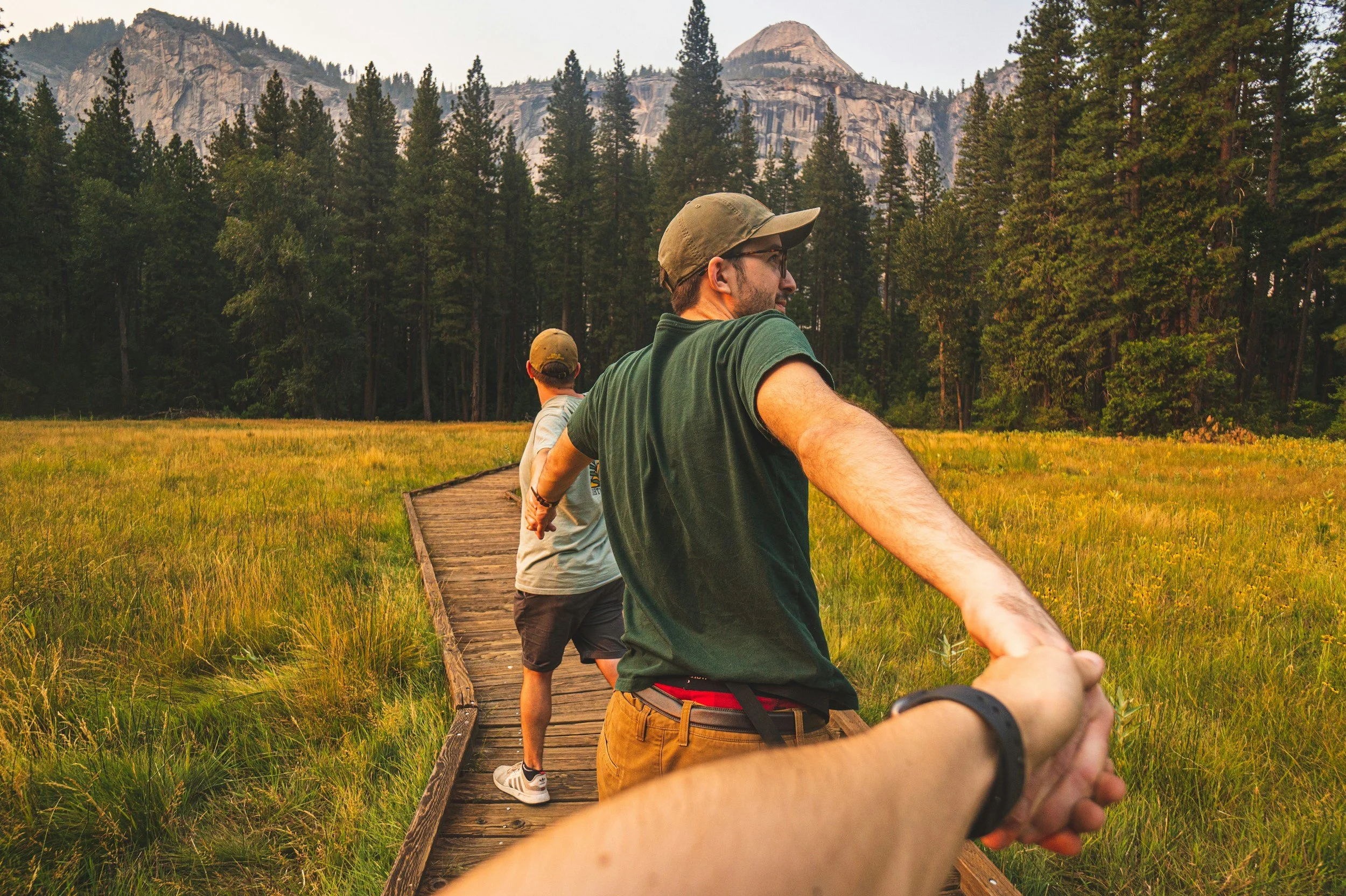 Three people holding hands in a line on a wooden path through a grassy field with a forest and mountains in the background.