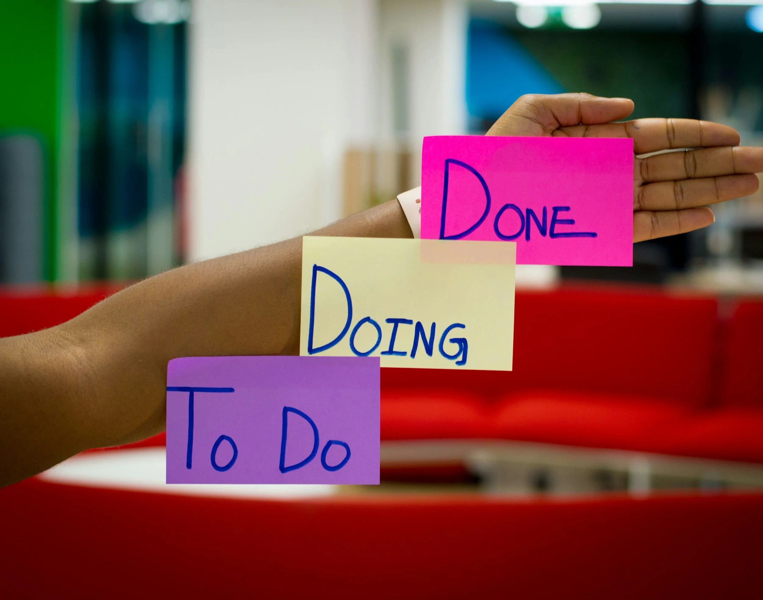 A person holding four colorful sticky notes arranged in a stepped manner, with the words 'To Do', 'Doing', and 'Done' written on them, indicating a task progress board.