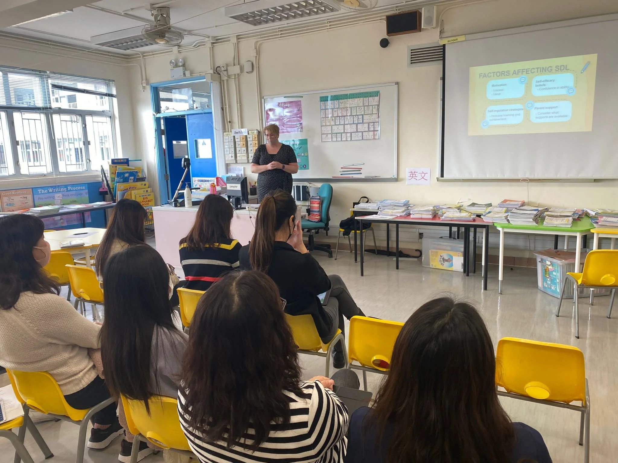 A classroom with several students sitting on yellow chairs, listening to a teacher standing at the front near a whiteboard and a projector screen displaying a presentation titled 'Factors Affecting SDL'. The room has large windows, shelves with books, and stacks of papers and books on tables.