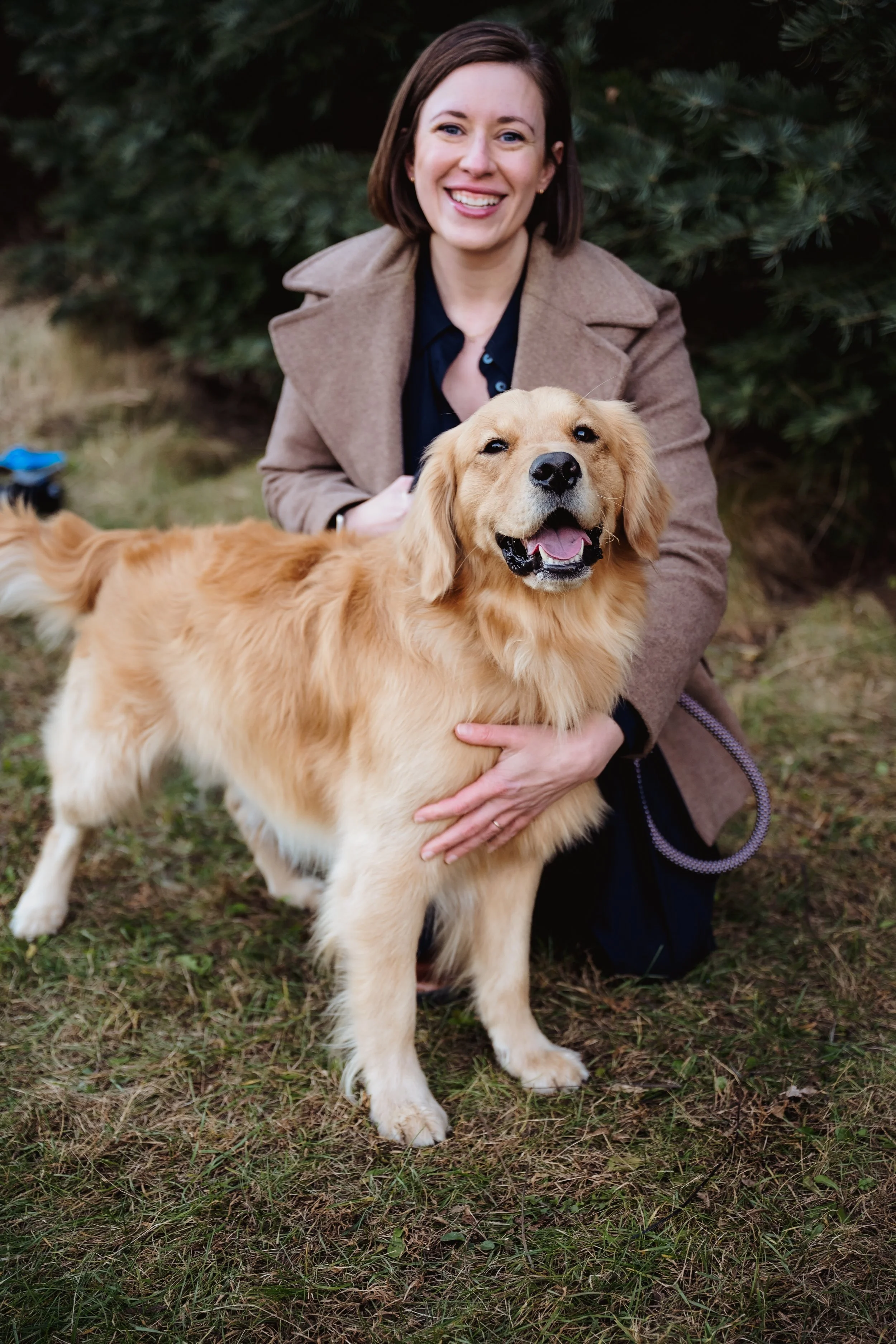 A woman smiling and kneeling on grass, holding a golden retriever dog with a happy expression, outdoors in front of pine trees.