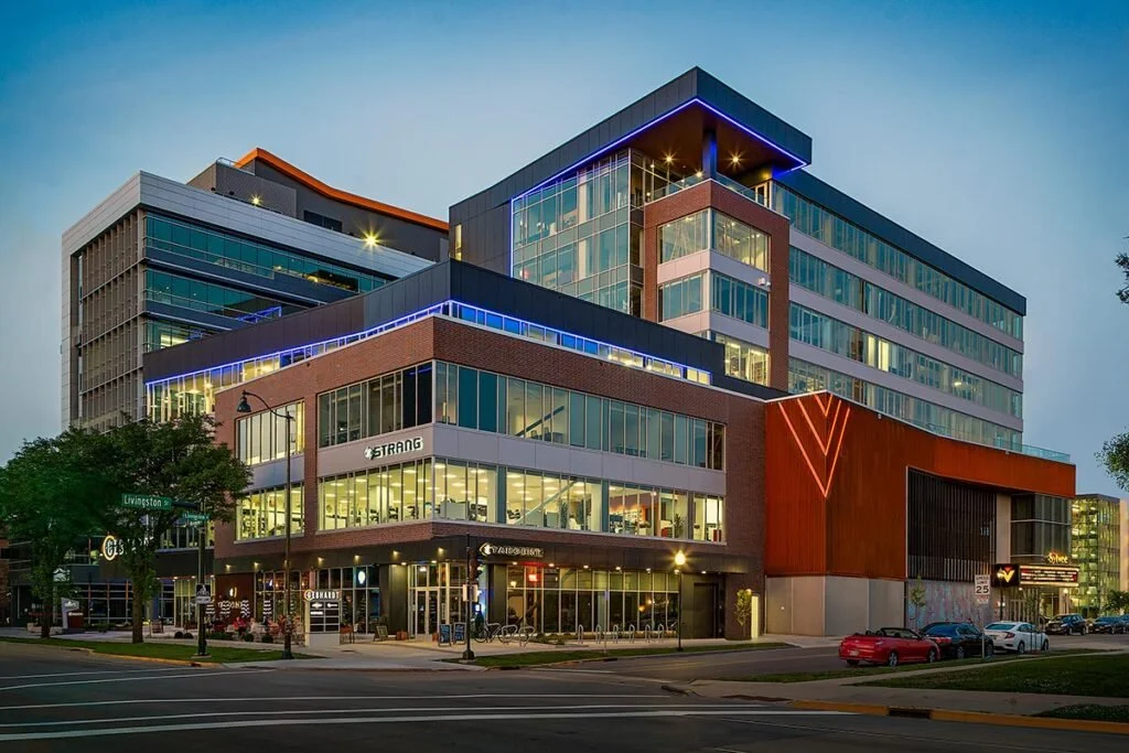 Modern multi-story office building with glass windows, illuminated with blue and neon accents, located on a city street at dusk.
