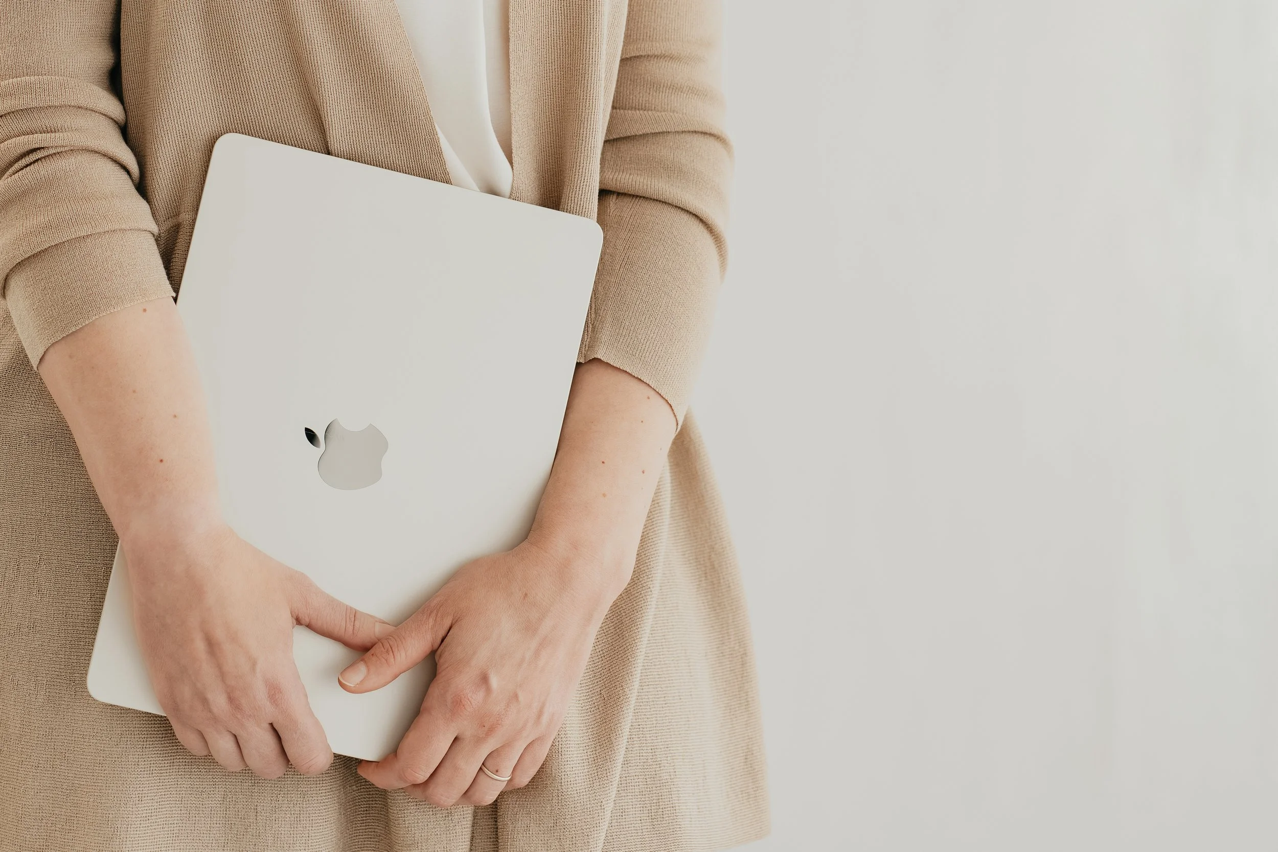 Person holding a closed silver MacBook laptop with a visible Apple logo on their front in front of a beige coat and neutral background.