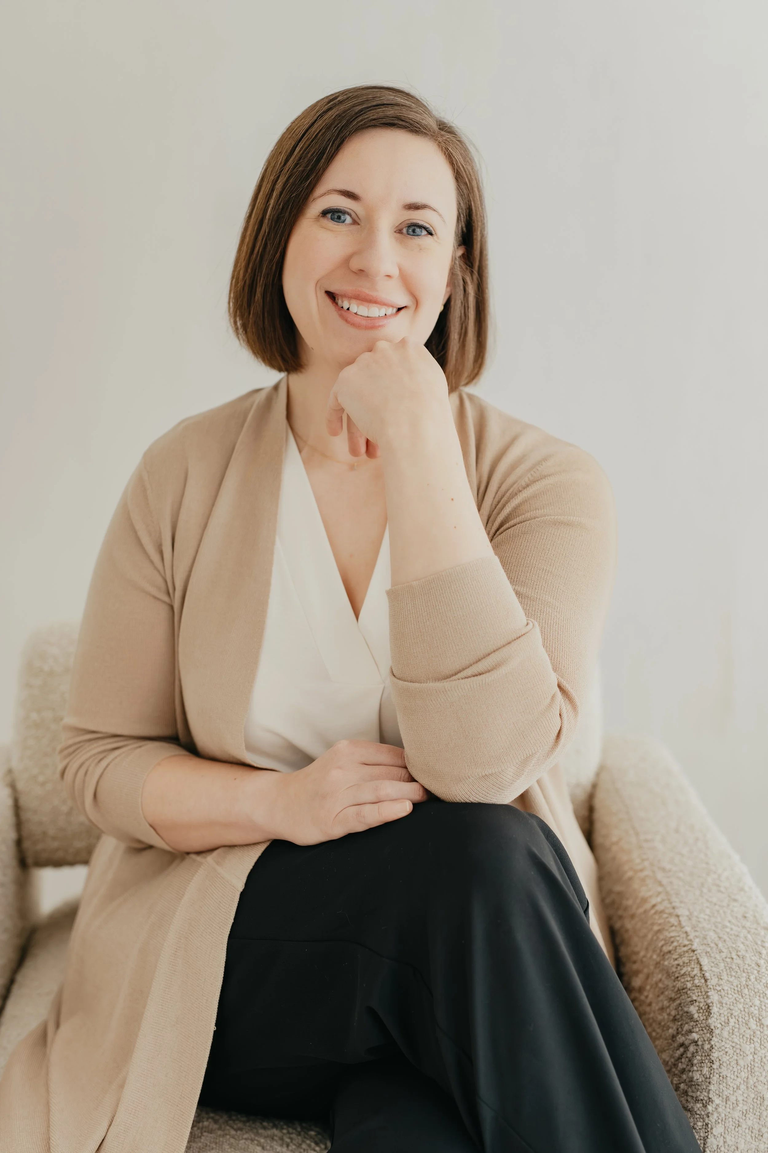 A woman with shoulder-length brown hair and blue eyes smiling while sitting on a beige textured armchair, wearing a beige cardigan over a white blouse and black pants, with her hand resting on her chin.