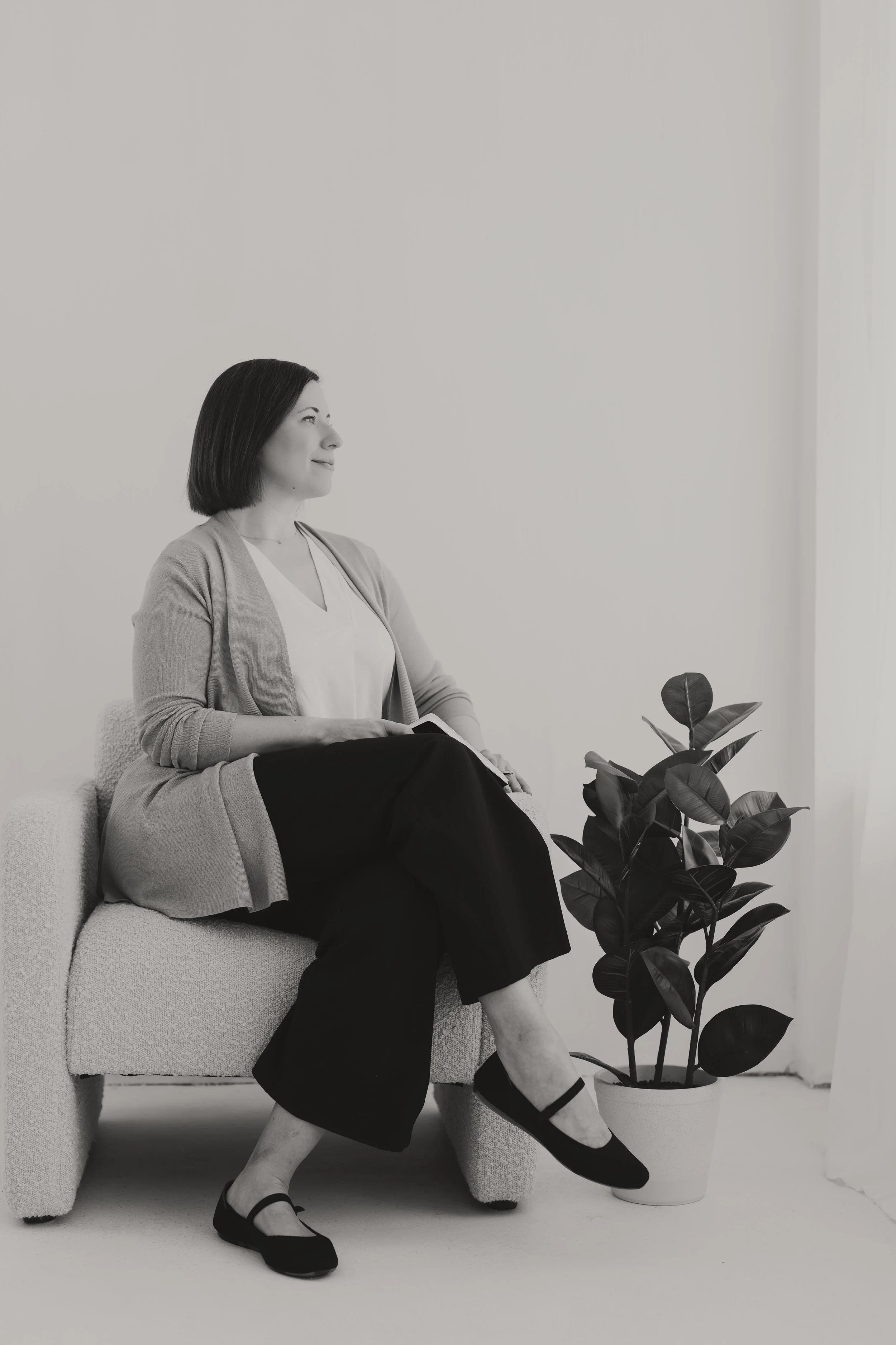 A woman with dark hair in a bob haircut sitting on a light-colored sofa, looking to her left, with a potted houseplant next to her, in a minimalist room with a plain wall and a sheer curtain.