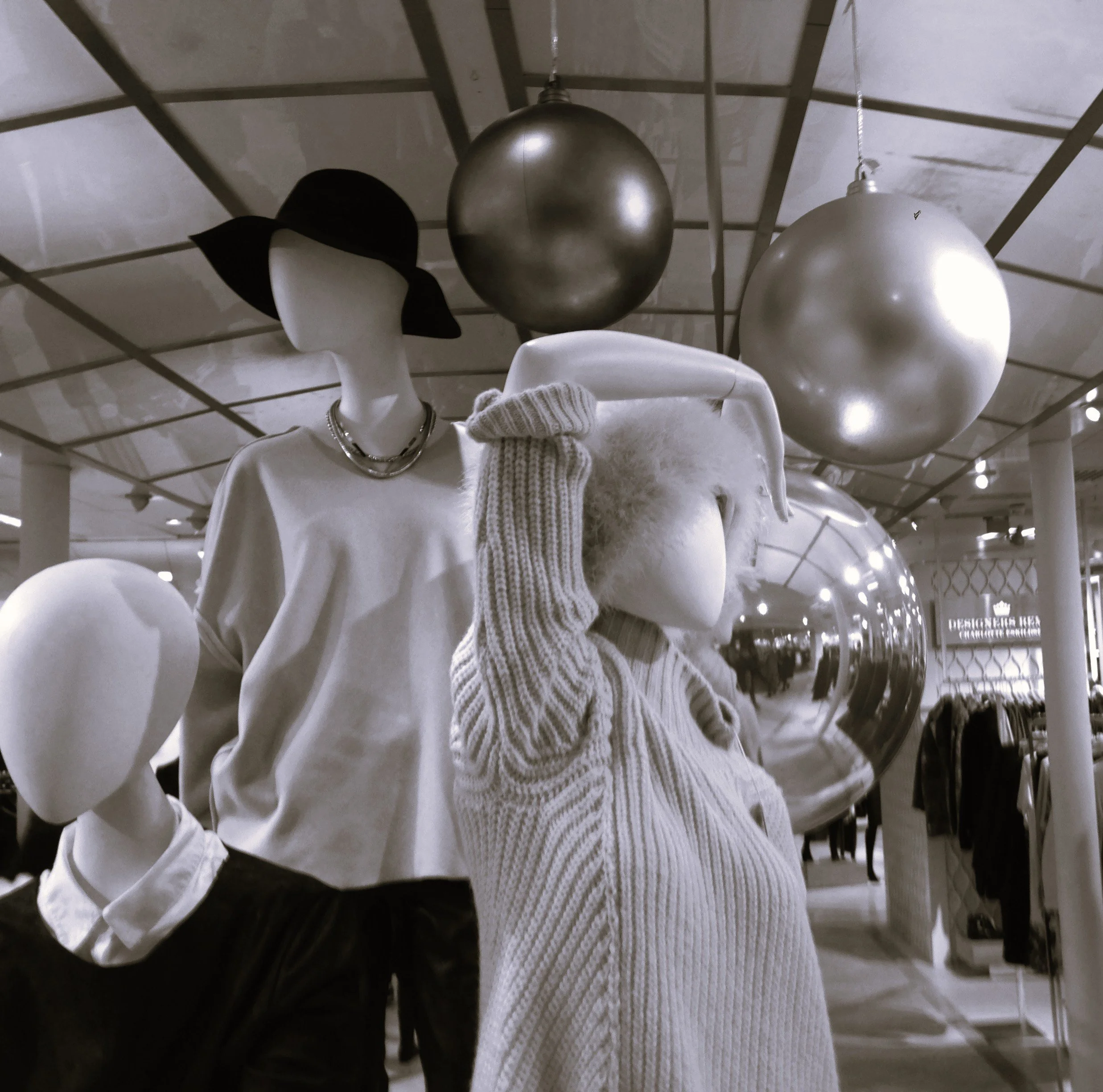Mannequins dressed in white garments with accessories, one wearing a black wide-brimmed hat and layered necklaces, inside a retail store with metallic holiday decorations hanging from the ceiling.