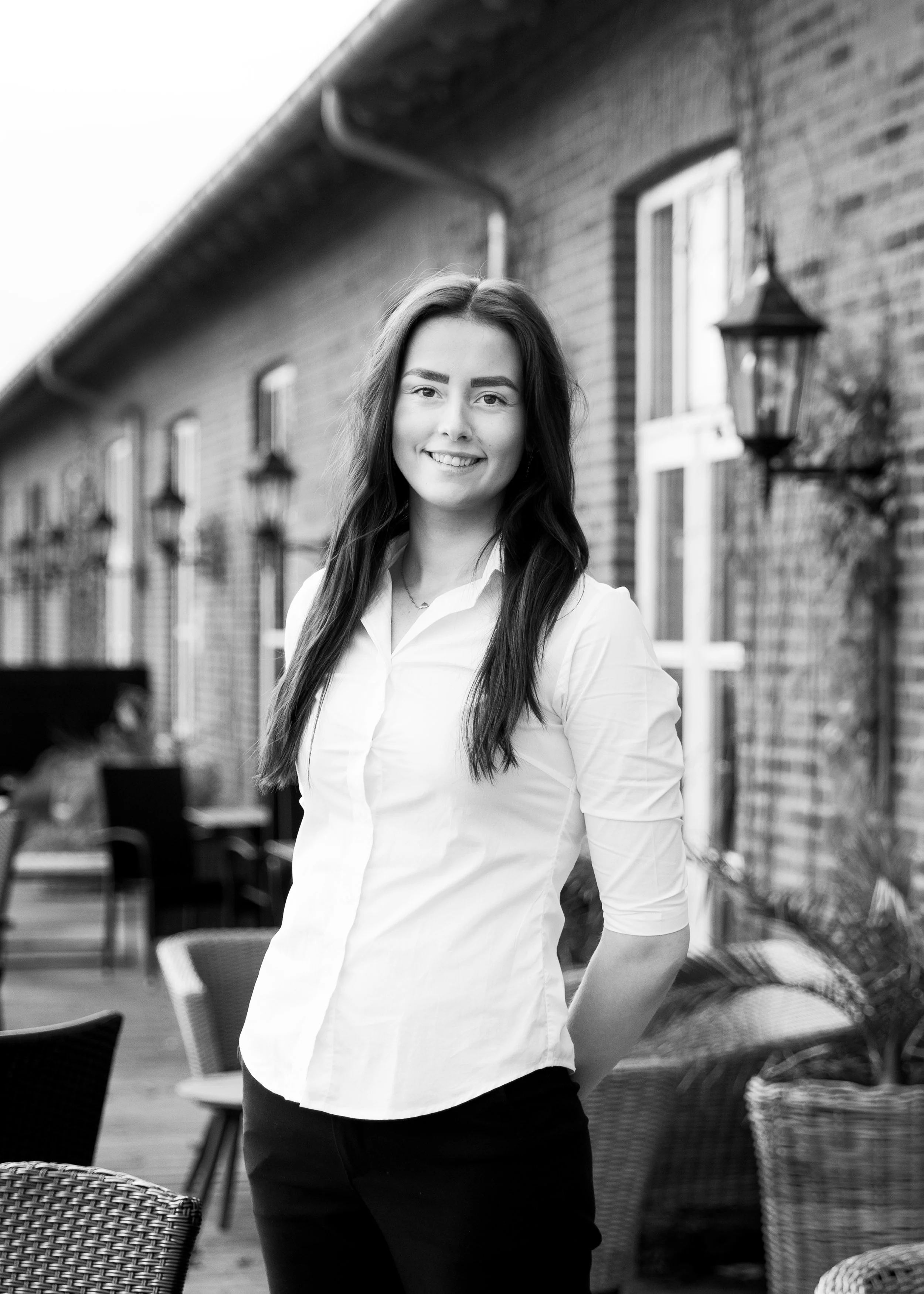 A young woman with long dark hair, wearing a white button-up shirt and dark pants, smiling while standing outdoors in front of a brick building with windows and lantern-style light fixtures.