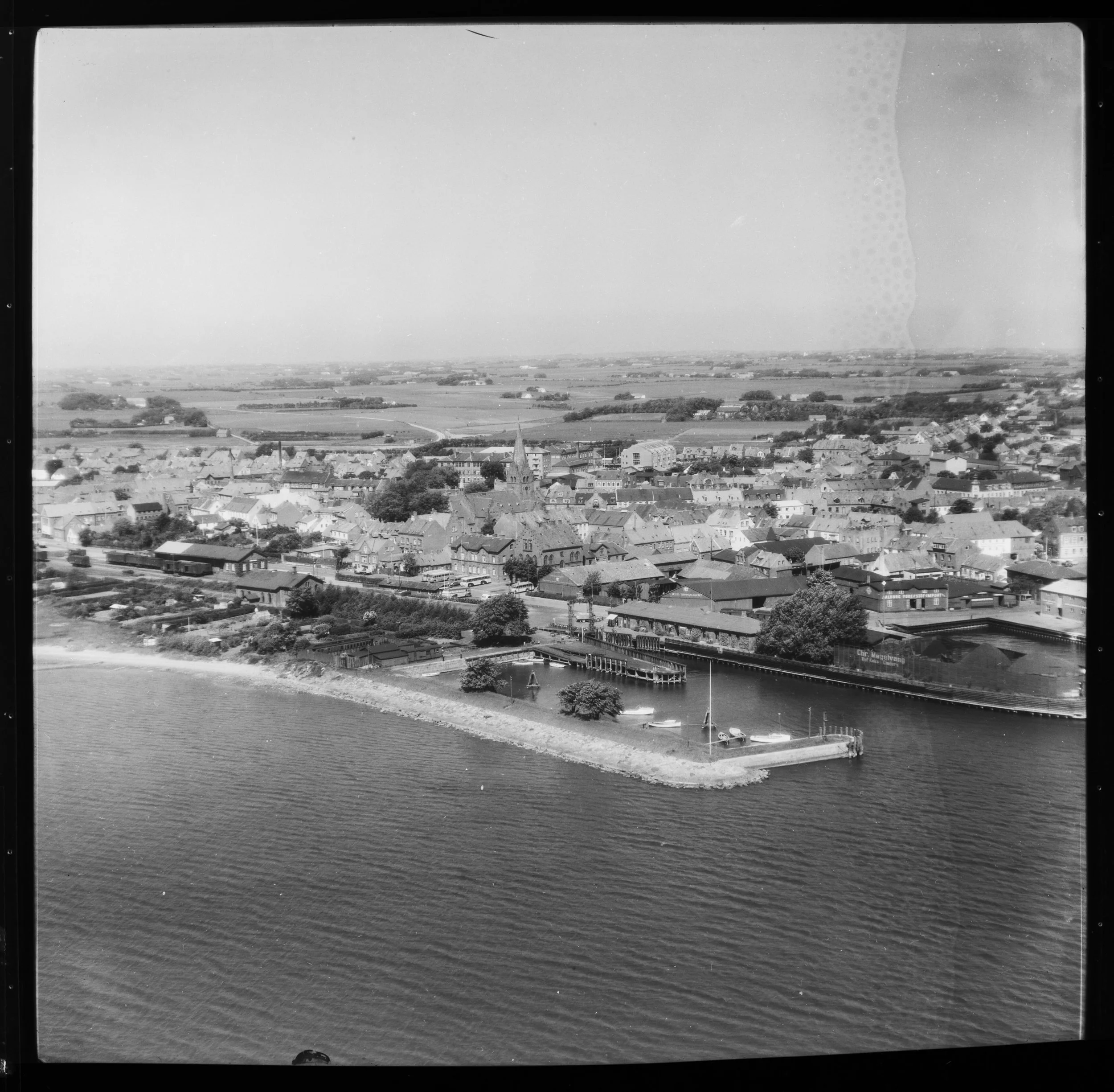 Sort/hvidt foto af en by med en lang bygningslinie langs en flod, der har en mole med små både. Ud over byen er der landbrugsområder med marker og åbne landskaber.