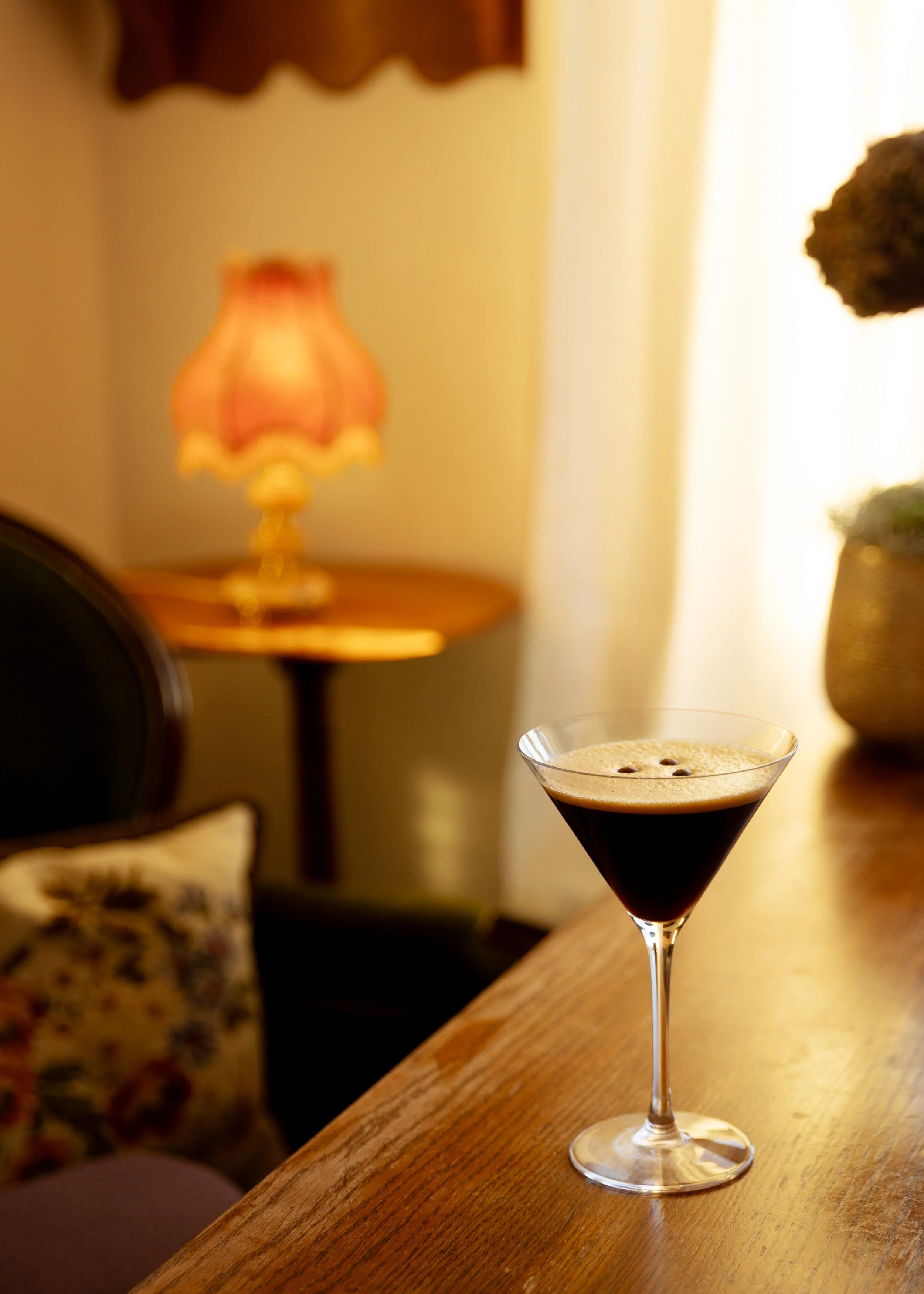 A cocktail glass filled espresso martini, topped with a foamy layer, placed on a wooden table in a warmly lit room.