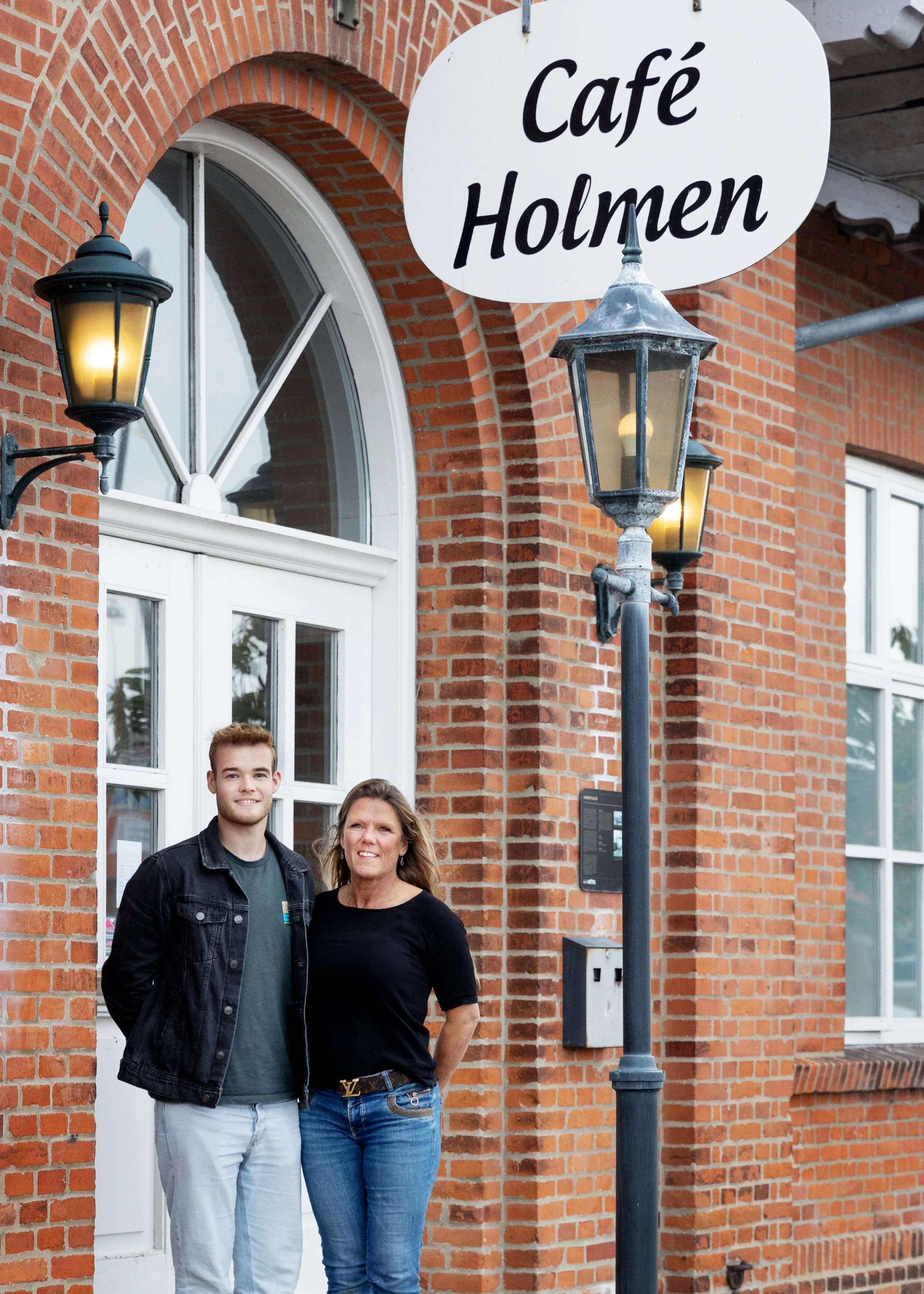 Two people, a young man and a woman, standing outside a brick building with a sign that reads 'Café Holmen' and vintage street lamps.