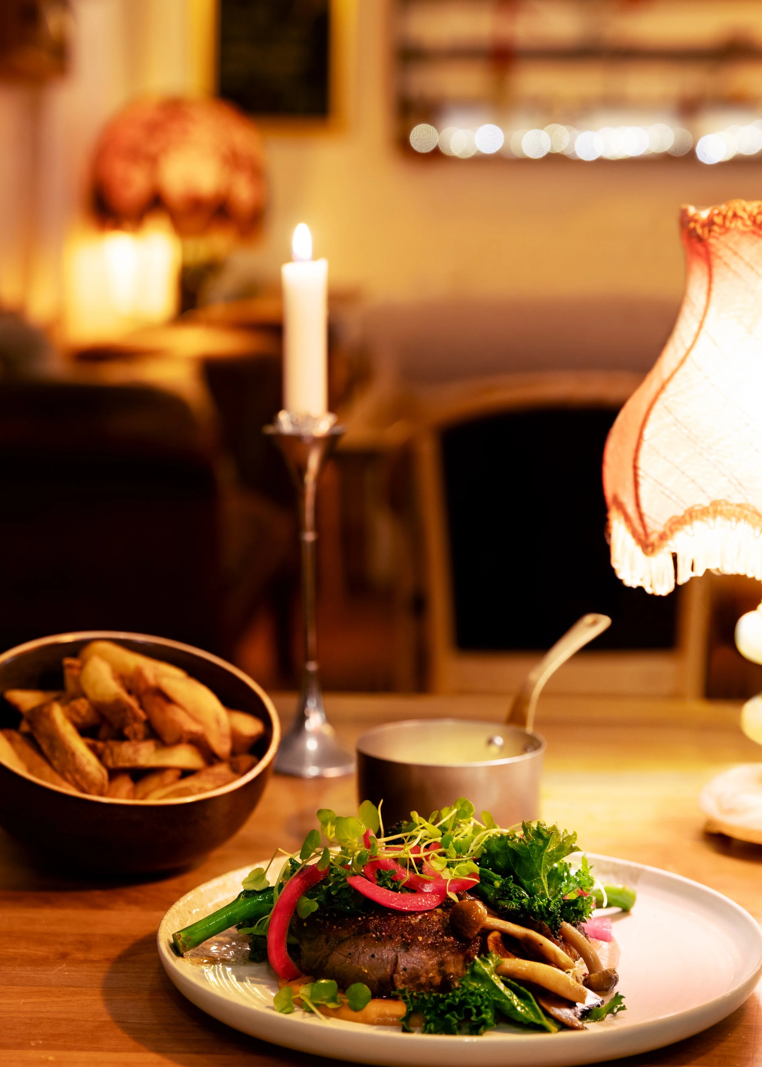 A dinner plate with cooked meat topped with vegetables and herbs, set on a wooden table with a bowl of fries, a candle, and a small pot of sauce in a cozy, warmly lit restaurant.