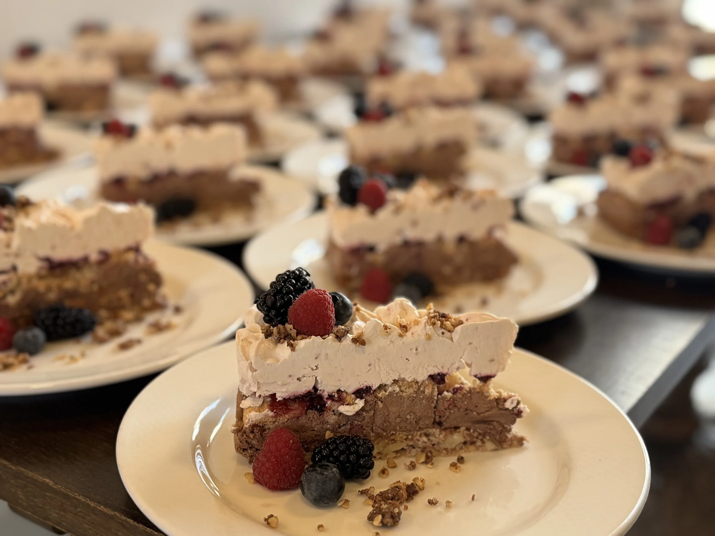 En serveret skive af lagkage med lag af chokolade og frugt, pyntet med bær og nødder, på en hvid tallerken, med flere skiver af lagkage i baggrunden.