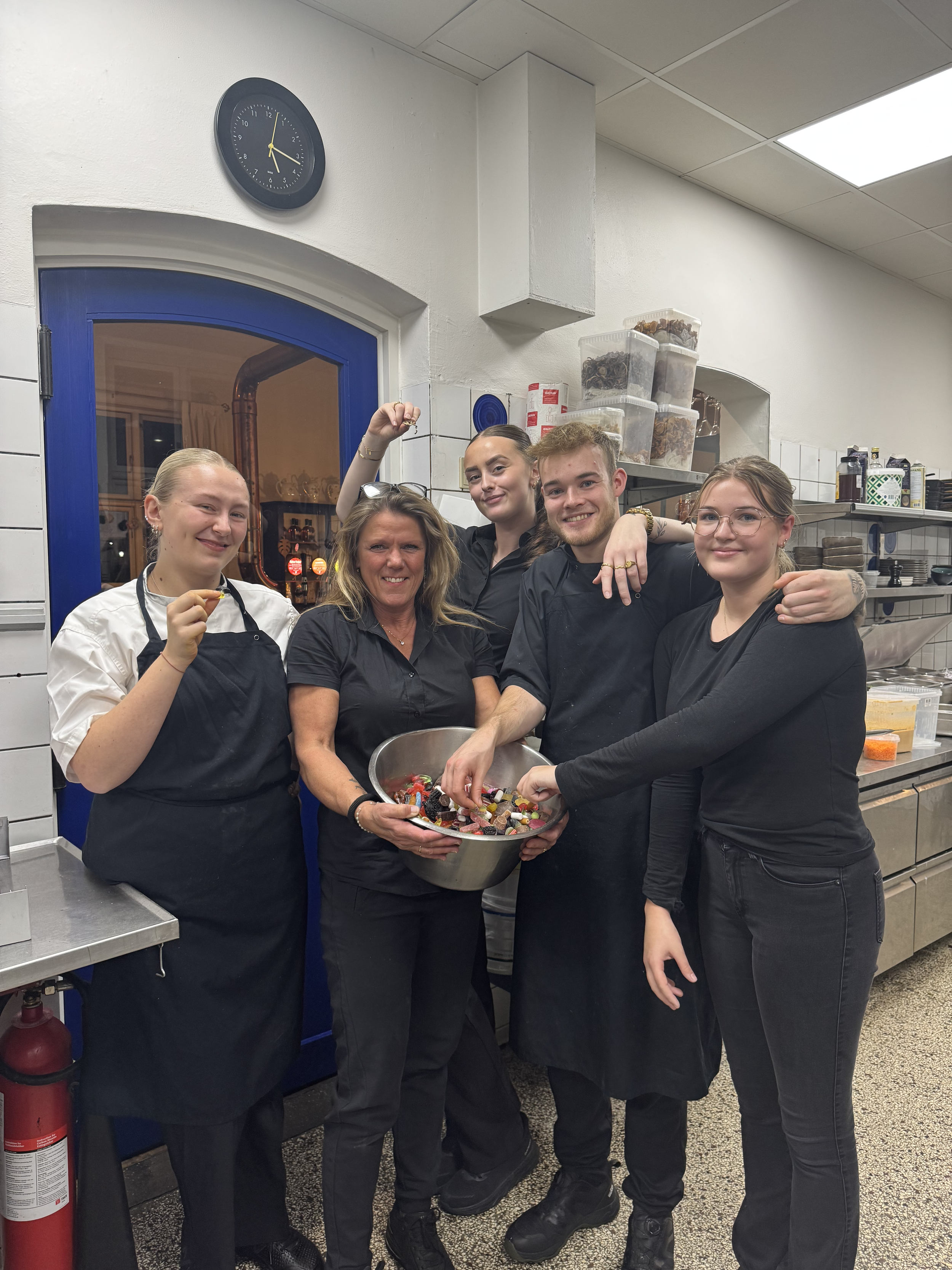 Fem mennesker i køkken, to kvinder og tre unge, alle iført sorte forklæder, står sammen med skål fyldt med slik og smiler.