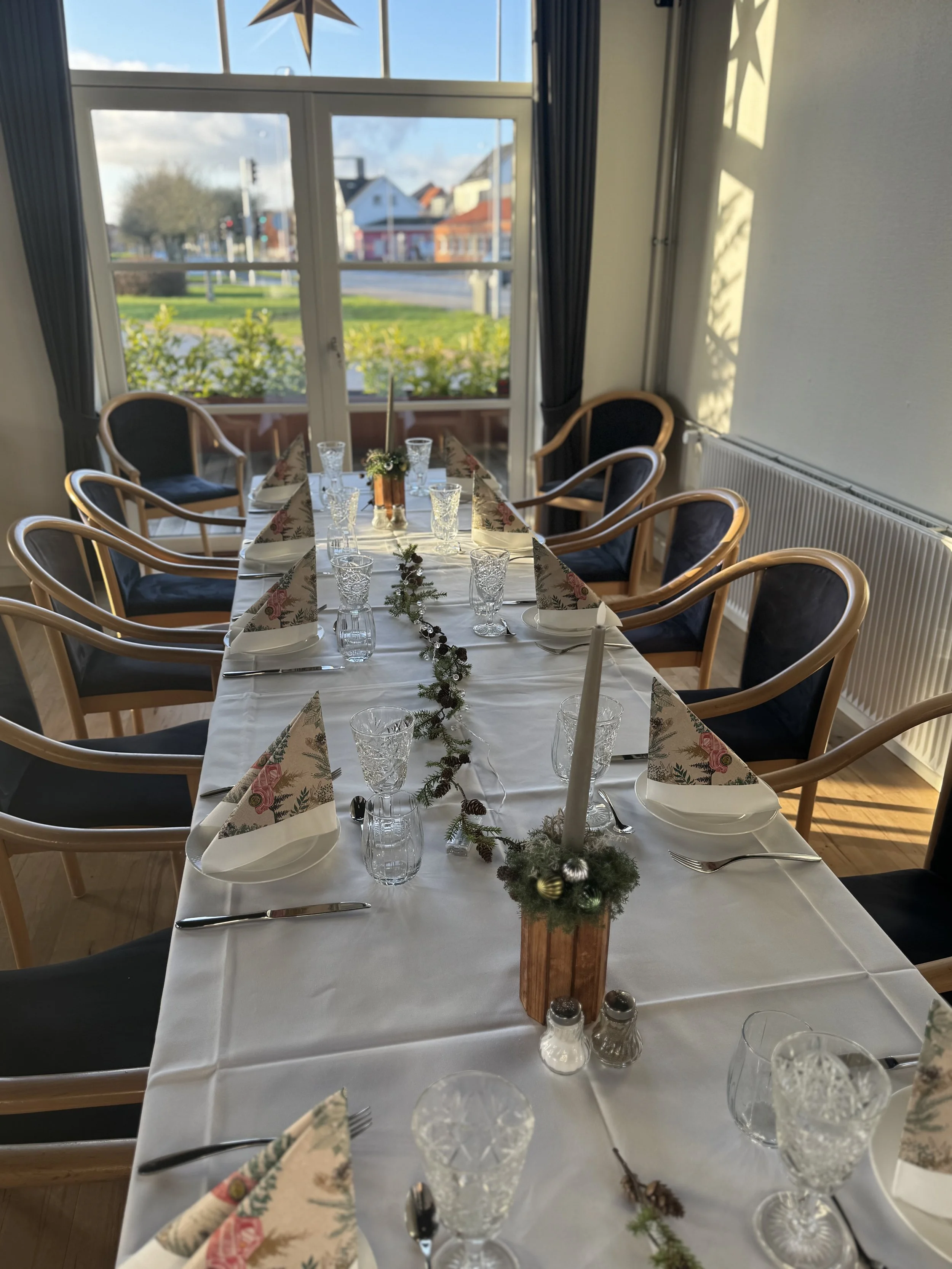 Indretning til jule middag med pynt, glas, bestik og dækkede tallerkener på et langbord.