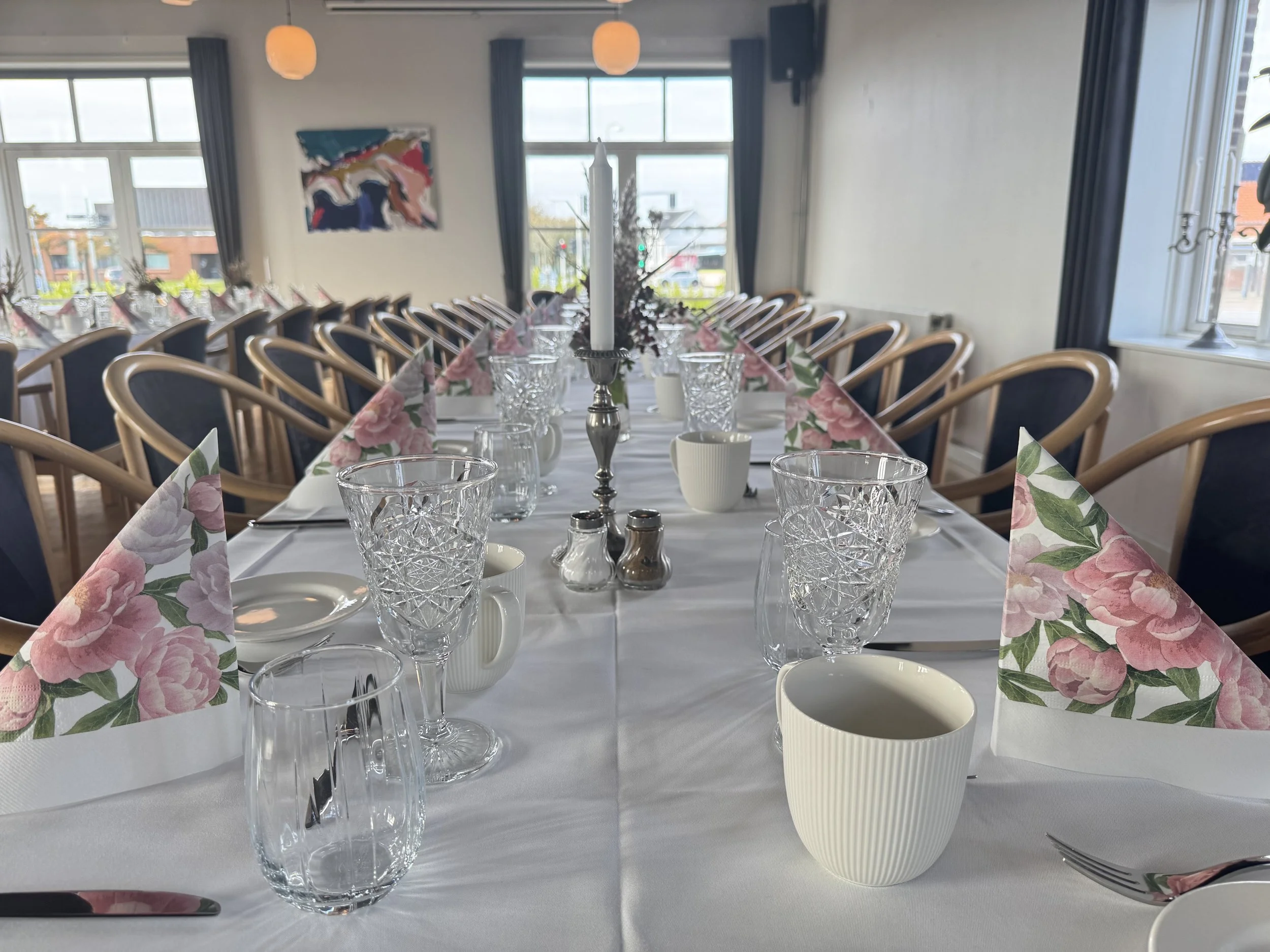 A long dining table set for a formal meal with wine glasses, napkins folded into triangle shapes with pink floral patterns, and white cups, in a well-lit room with large windows and wooden chairs.