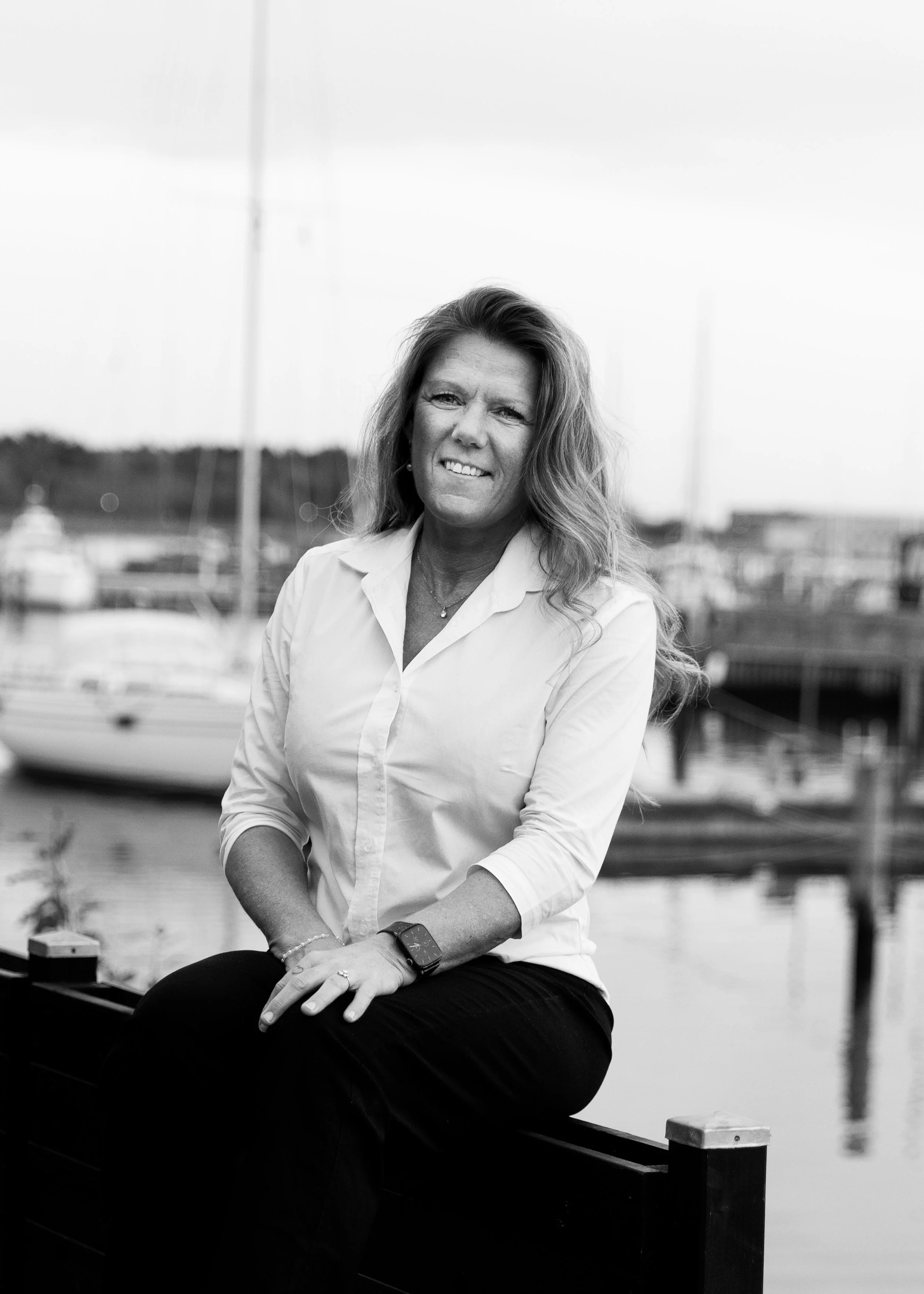 A woman with long wavy hair sitting on a bench at a marina, with sailboats and water in the background, captured in black and white.