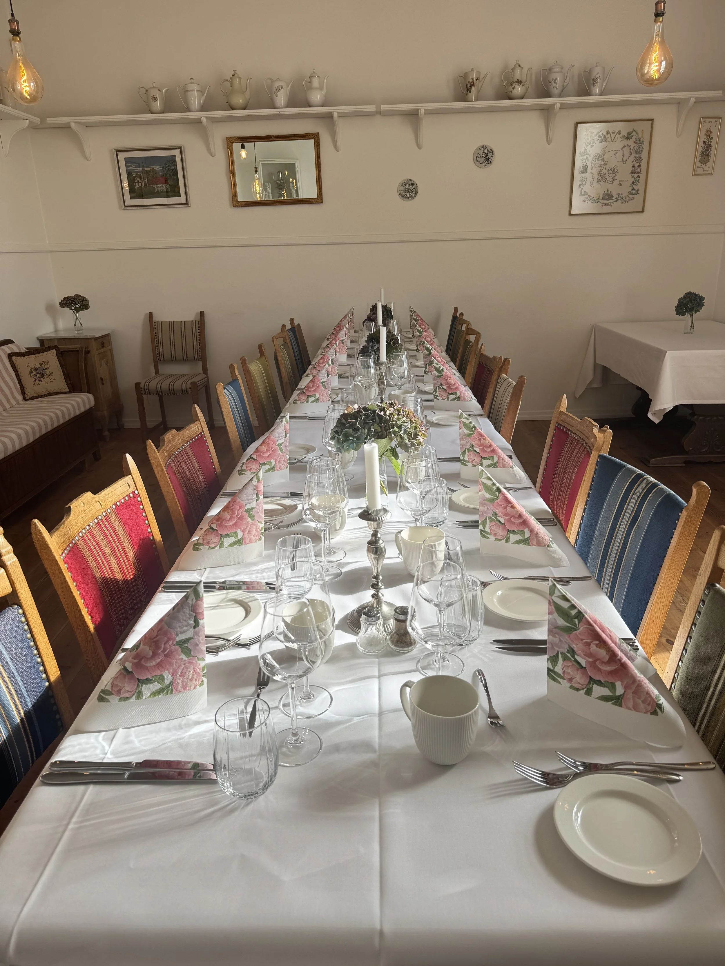 Bord dækket til en festmiddag med blomsterdekorationer, glas, tallerkener og bestik, omgivet af stole med forskellige farvede stoffer.