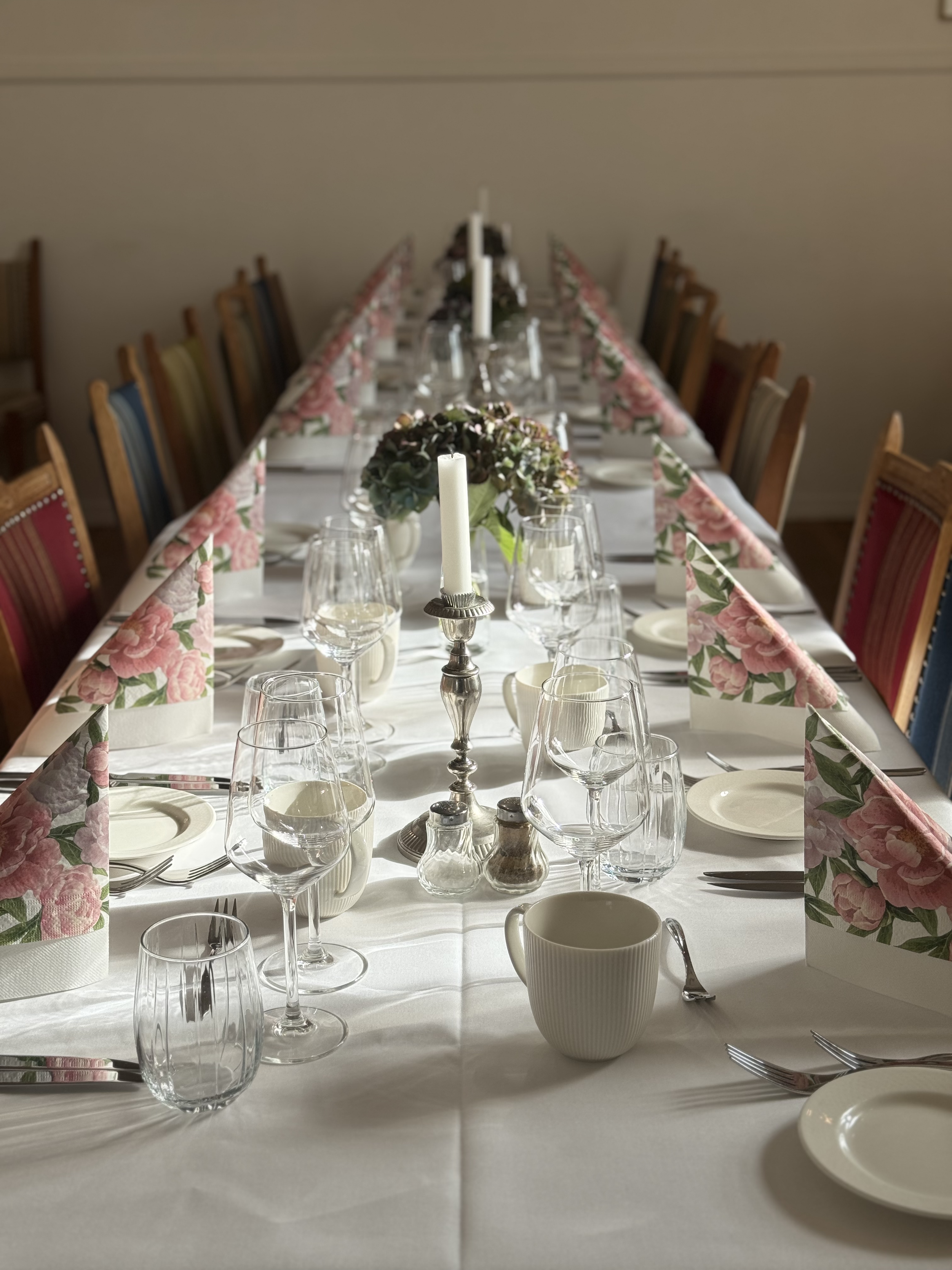 Langt spisebord dækket med hvid dug, med tallerkener, glas, bestik, blomsterarrangement og lyse lyspunkter.