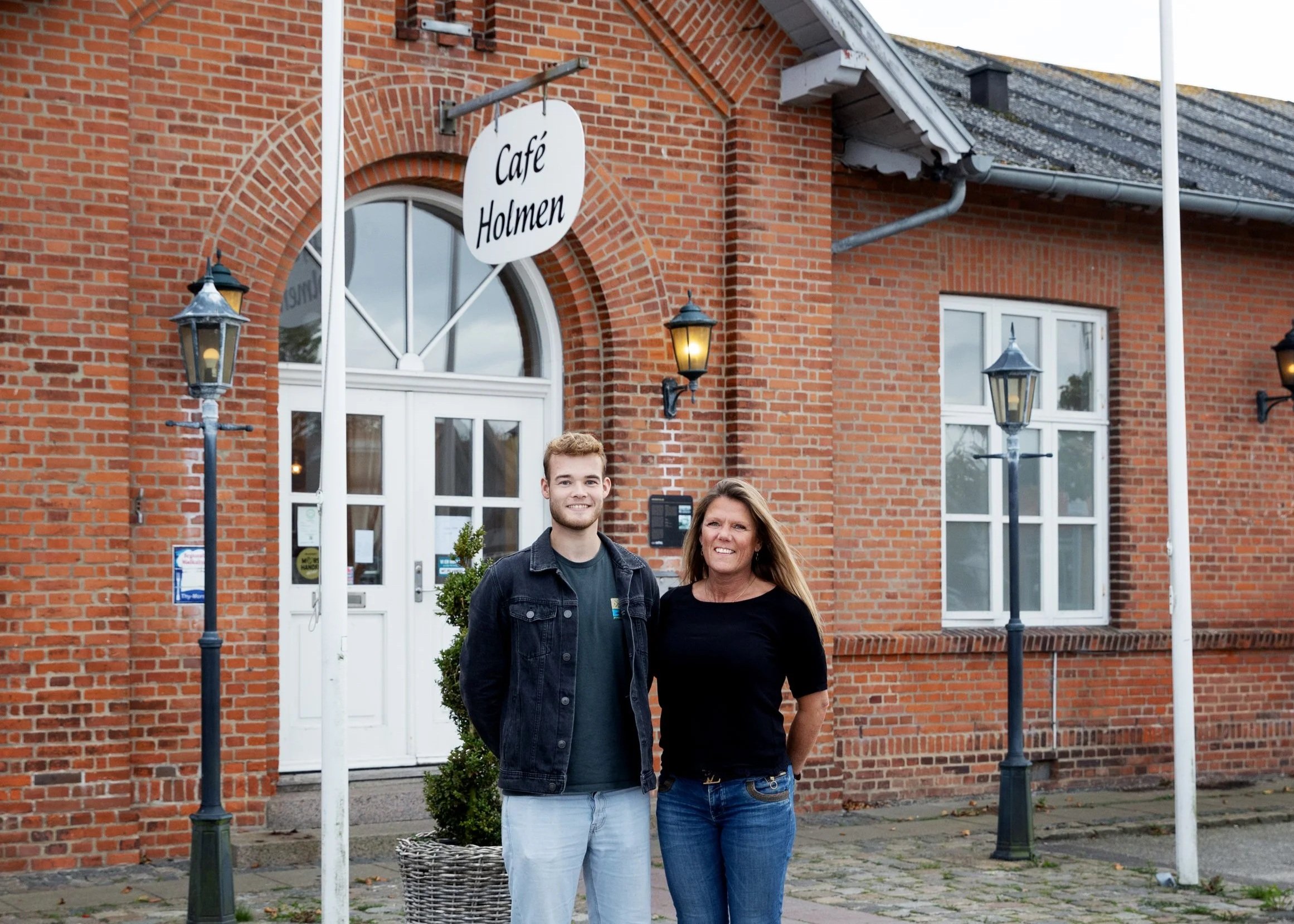 A young man and a woman standing outside of a red brick building with a sign reading 'Café Holmen'. The man is wearing a dark jacket and light pants, and the woman is wearing a black top and blue jeans. There are two street lamps and a potted plant near the entrance.