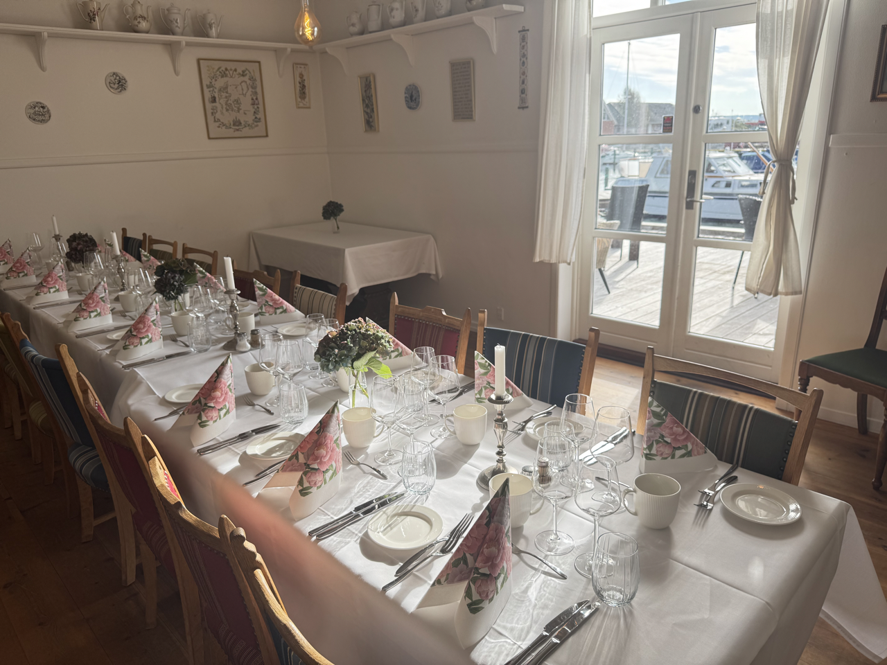 Et langt middagsbord dækket med hvide duge, rosenmønstrede servietter, glas, tallerkener, bestik, lysestager med levende lys, og blomsterdekorationer, i et lyst værelse med store vinduer, der vender ud mod en terrasse med udsigt over bådhamne.