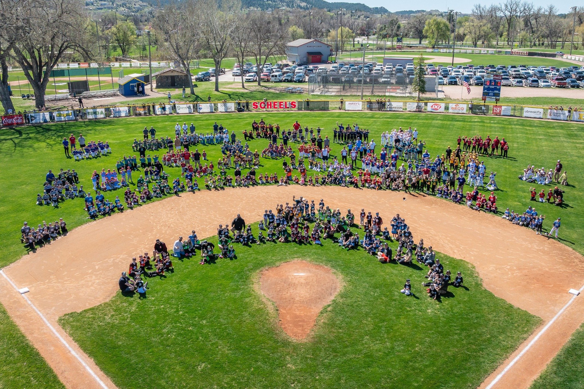 A large group of children and adults gathered on a baseball field for a group photo, with some dressed in sports uniforms and hats, under a sunny sky with trees and parking lot in the background.
