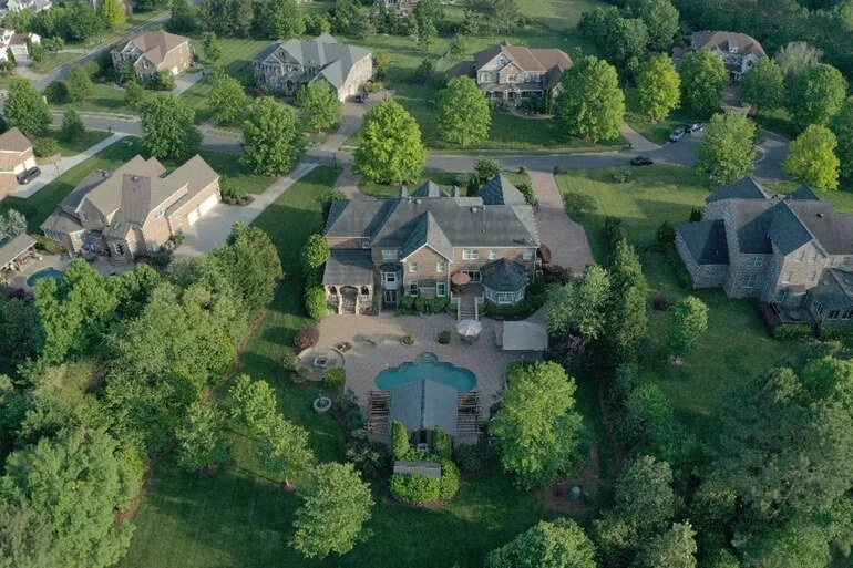 Aerial view of a suburban neighborhood featuring a large house with a backyard swimming pool, surrounded by green trees and other houses.