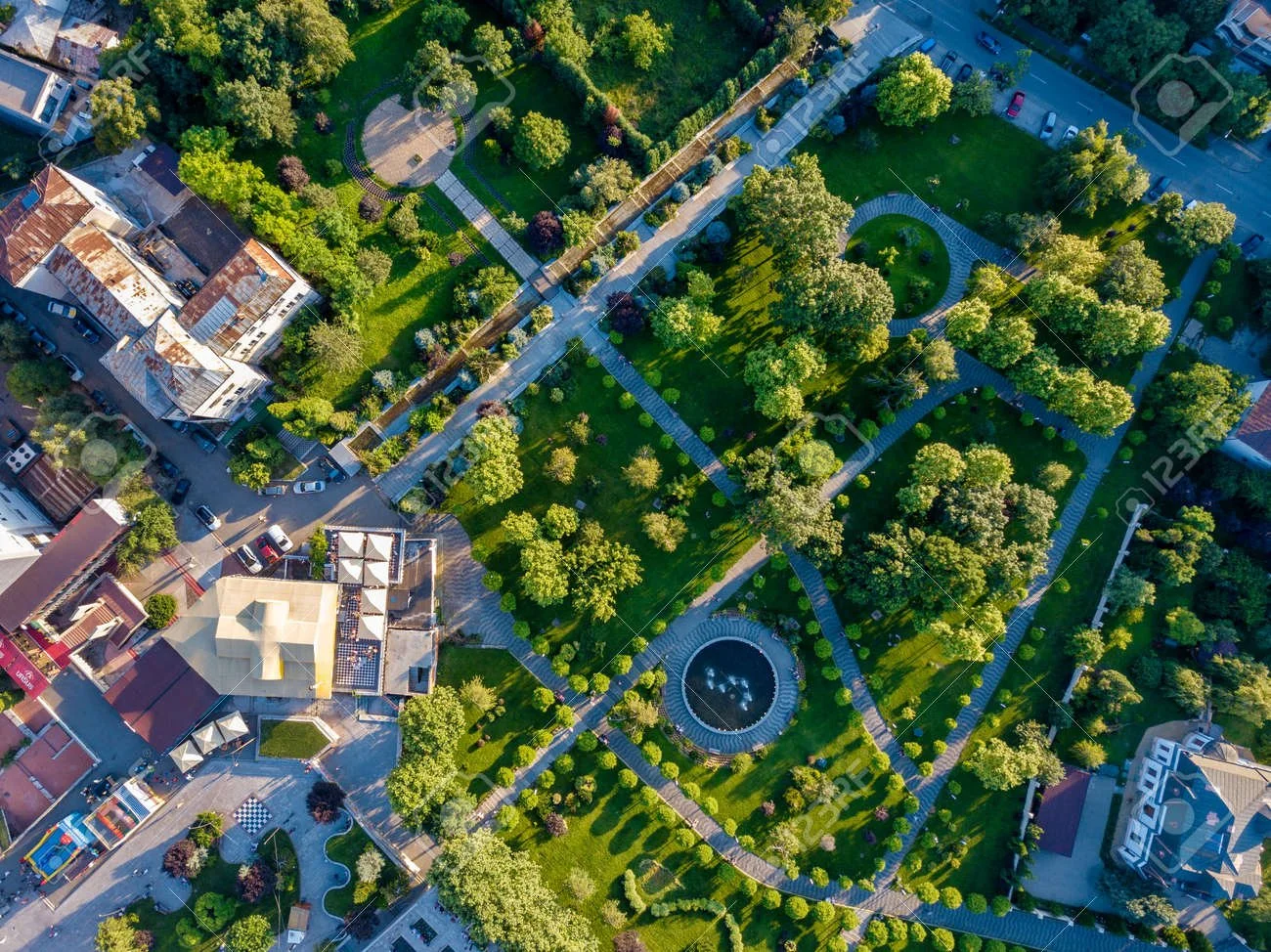Aerial view of an urban park with walking paths, green lawns, trees, a fountain, and surrounding buildings and parking lots.