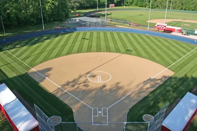 A baseball field with a dirt infield and grass outfield, surrounded by a blue running track, with trees and a small building in the background.