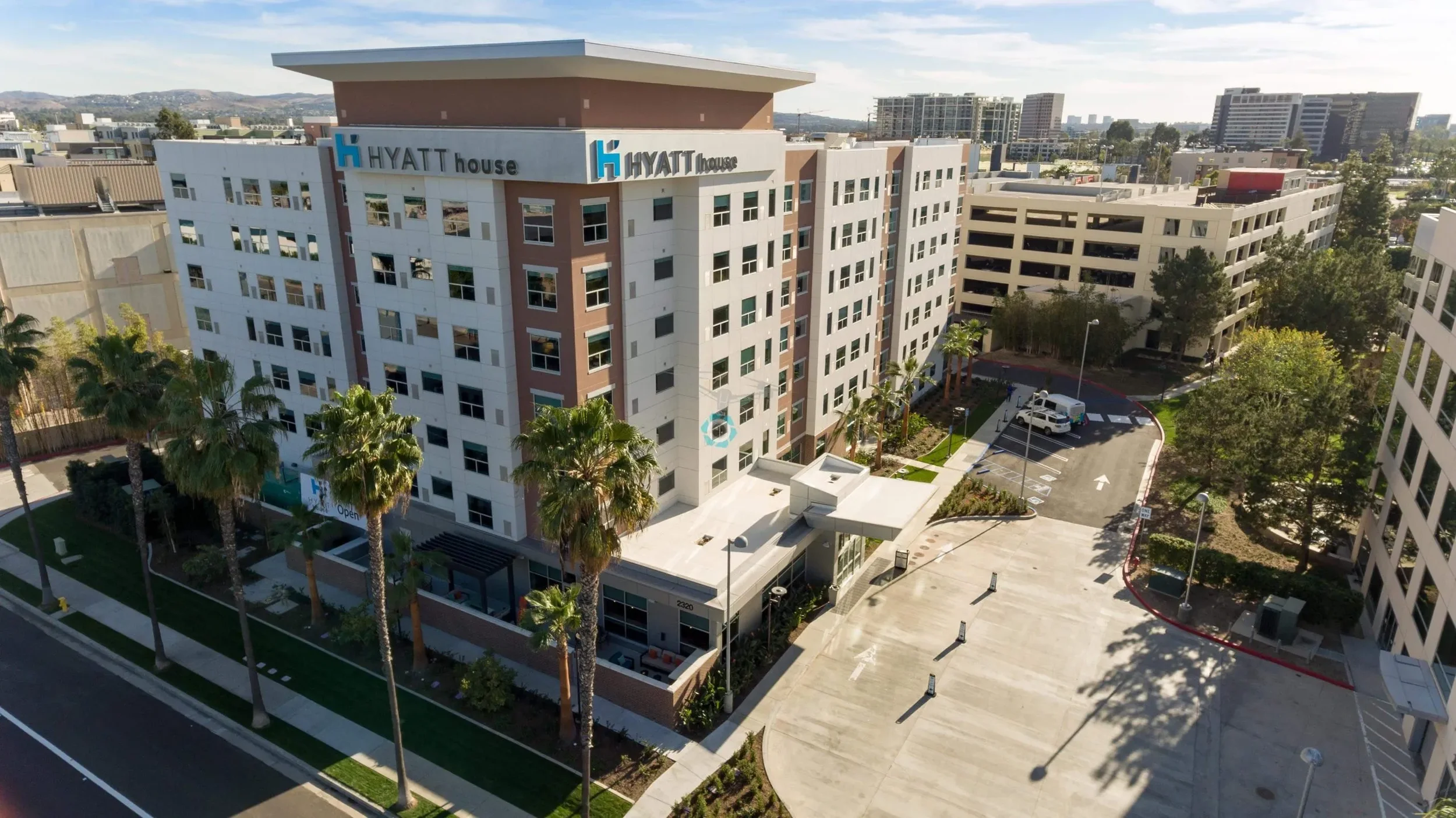 A modern multi-story hotel building labeled 'HYATT house' with a few palm trees outside, a parking area, and other nearby buildings in an urban setting, with mountains in the distance under a clear sky.