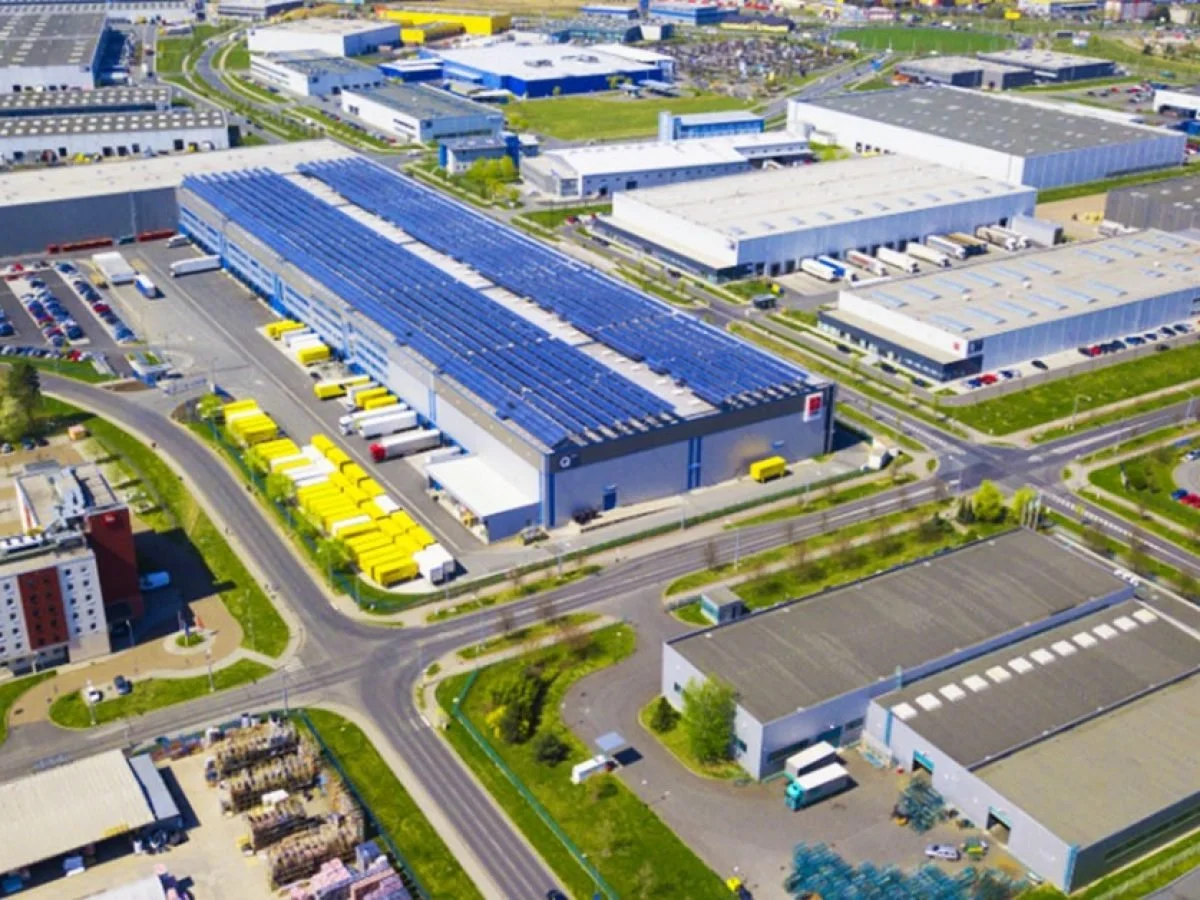 Aerial view of a large industrial warehouse with solar panels on the roof, surrounded by parking lots and other buildings.