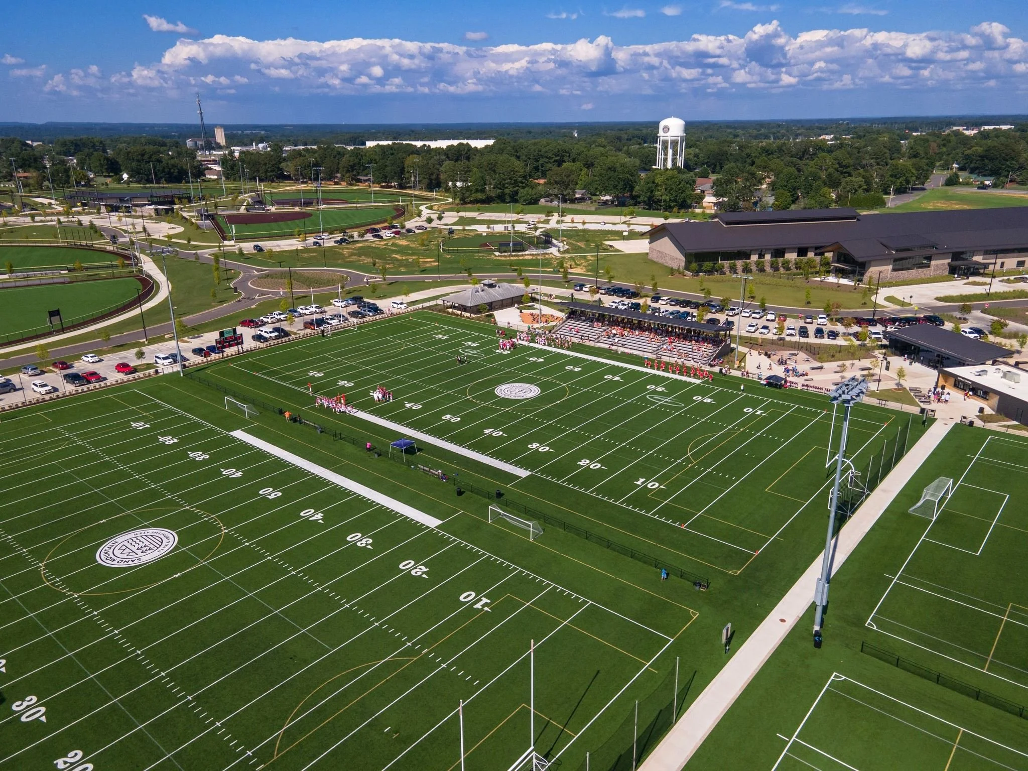 Aerial view of a football stadium with a game in progress, surrounded by parking lots, roads, and green fields, under a partly cloudy sky.