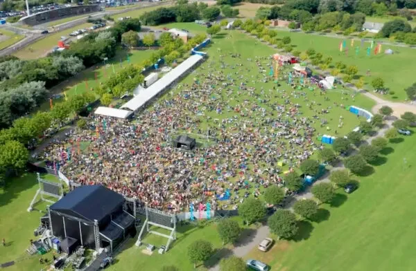Aerial view of a large outdoor concert with a big crowd on a grassy field, stage setup, tents, and trees surrounding the area.