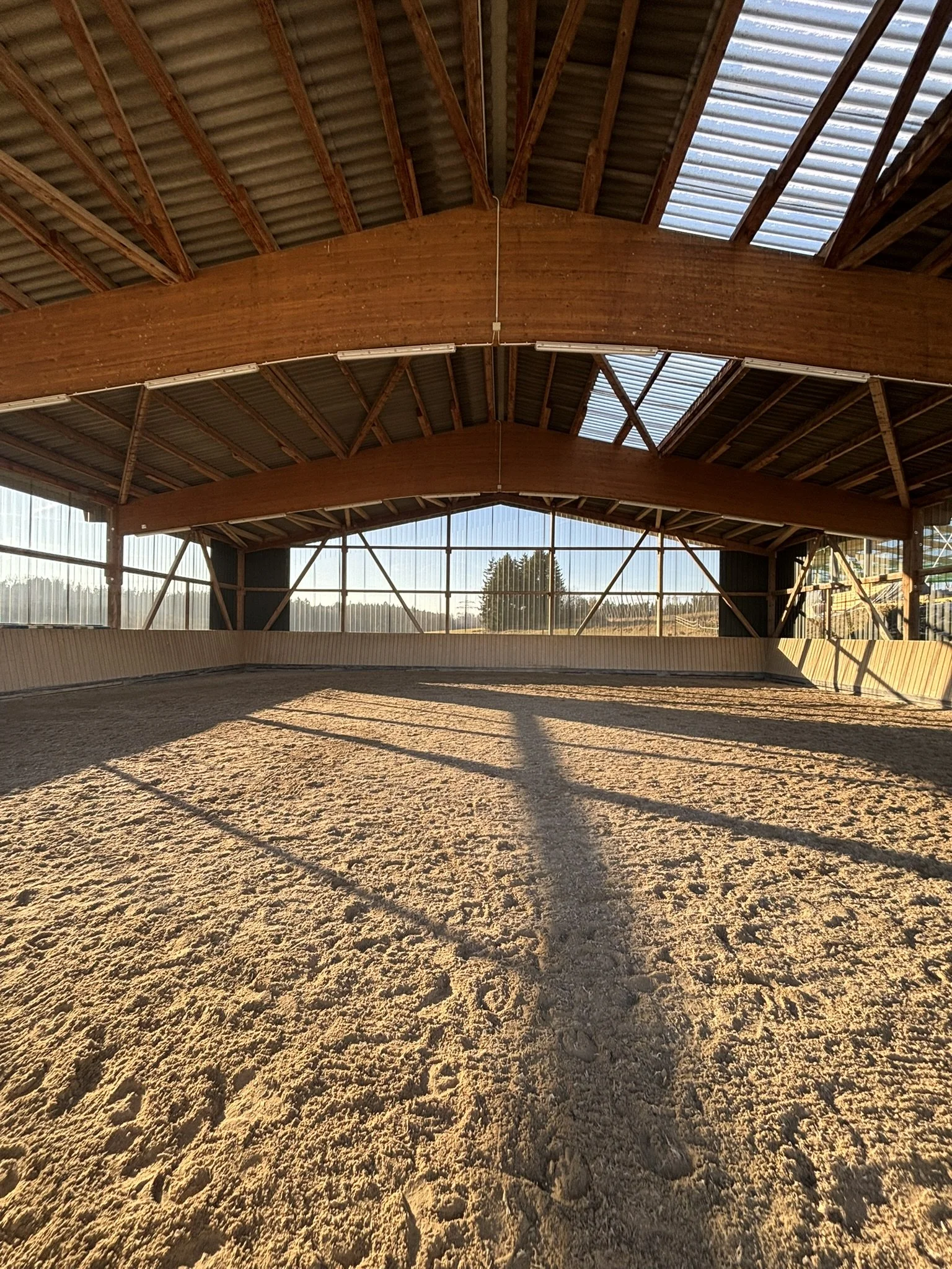 Innenansicht eines Reitstadions mit sandigem Boden und Holz- und Metallkonstruktion, Sonnenlicht wirft Schatten auf den Boden.