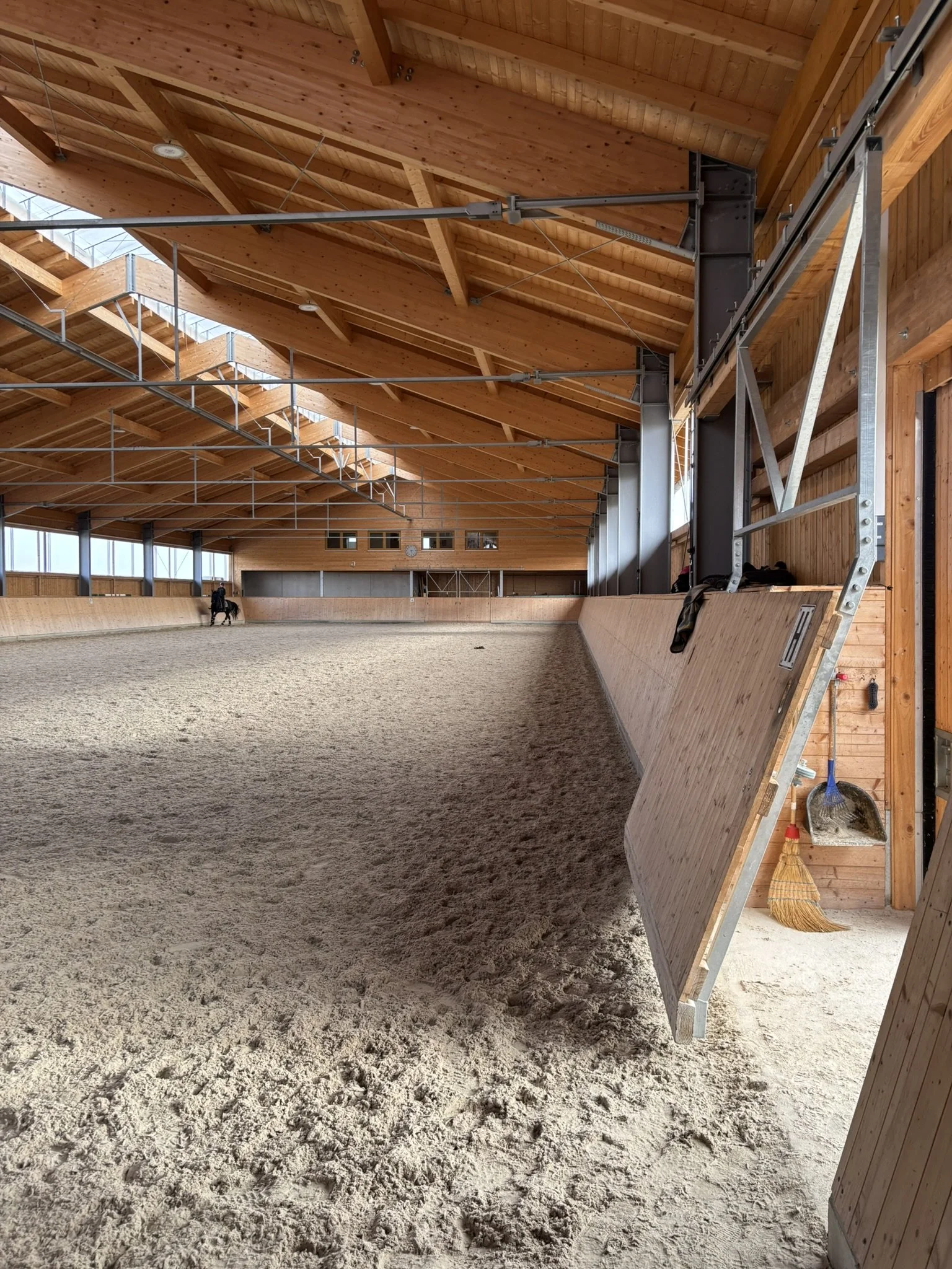 Innenansicht eines Reithalls mit Sandboden, Holzwänden und einer hohen Holzdecke. Es ist eine Person auf einem Pferd im Abstand sichtbar.
